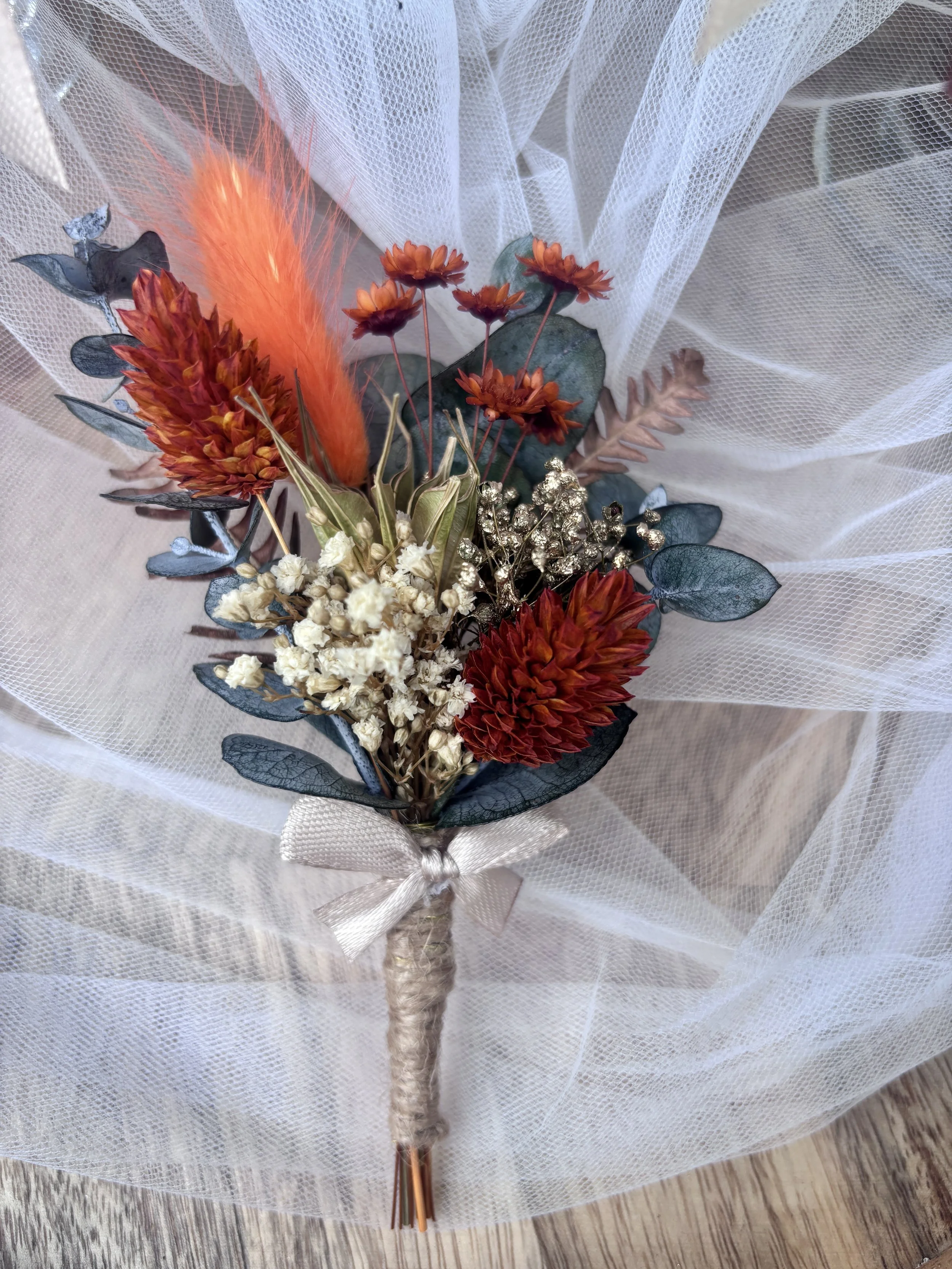 Boutonnière de marié en fleurs séchées et stabilisées, aux tons terracotta, orange, ivoire et eucalyptus, avec une touche dorée, idéale pour un mariage bohème.
