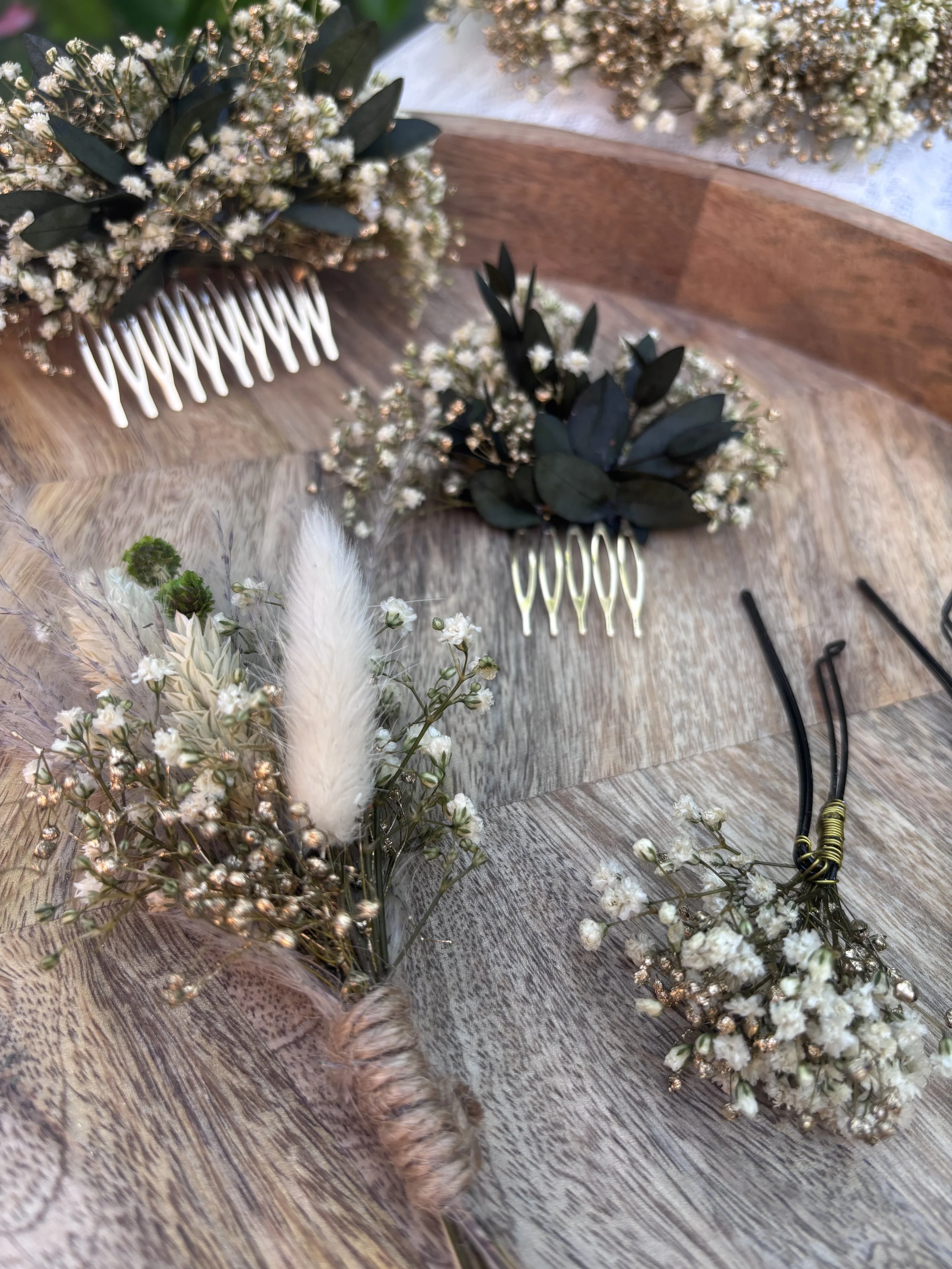 Une composition florale avec des fleurs blanches et des feuilles vertes, accompagnée d'une pince à cheveux dorée, posée sur une surface en bois.