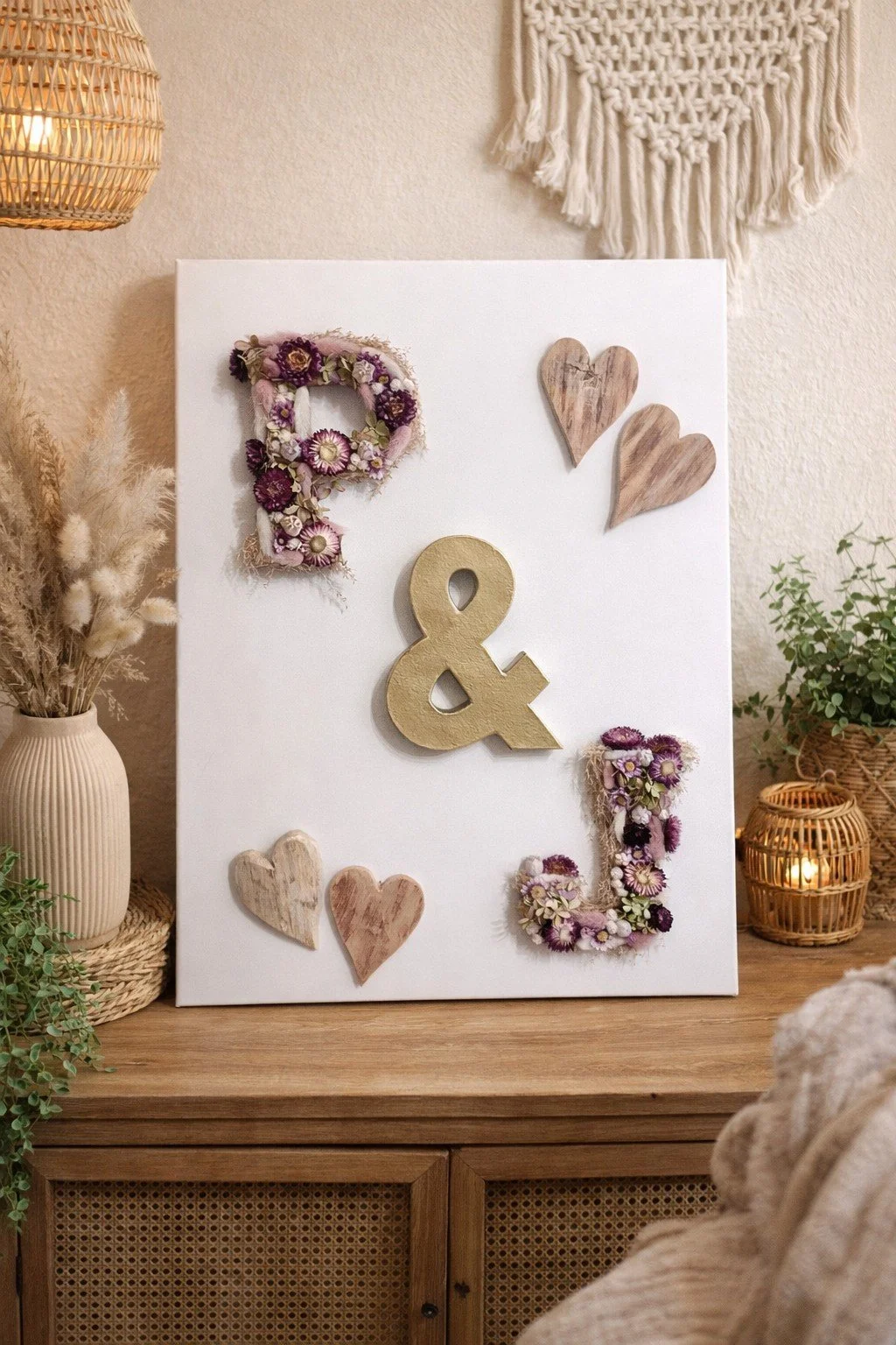 Décoration murale avec des lettres en bois et en fleurs, contenant un mélange de cœurs en bois et en fleurs, sur un fond blanc, dans un décor intérieur chaleureux avec une lampe en rotin, des plantes et une commode en bois.