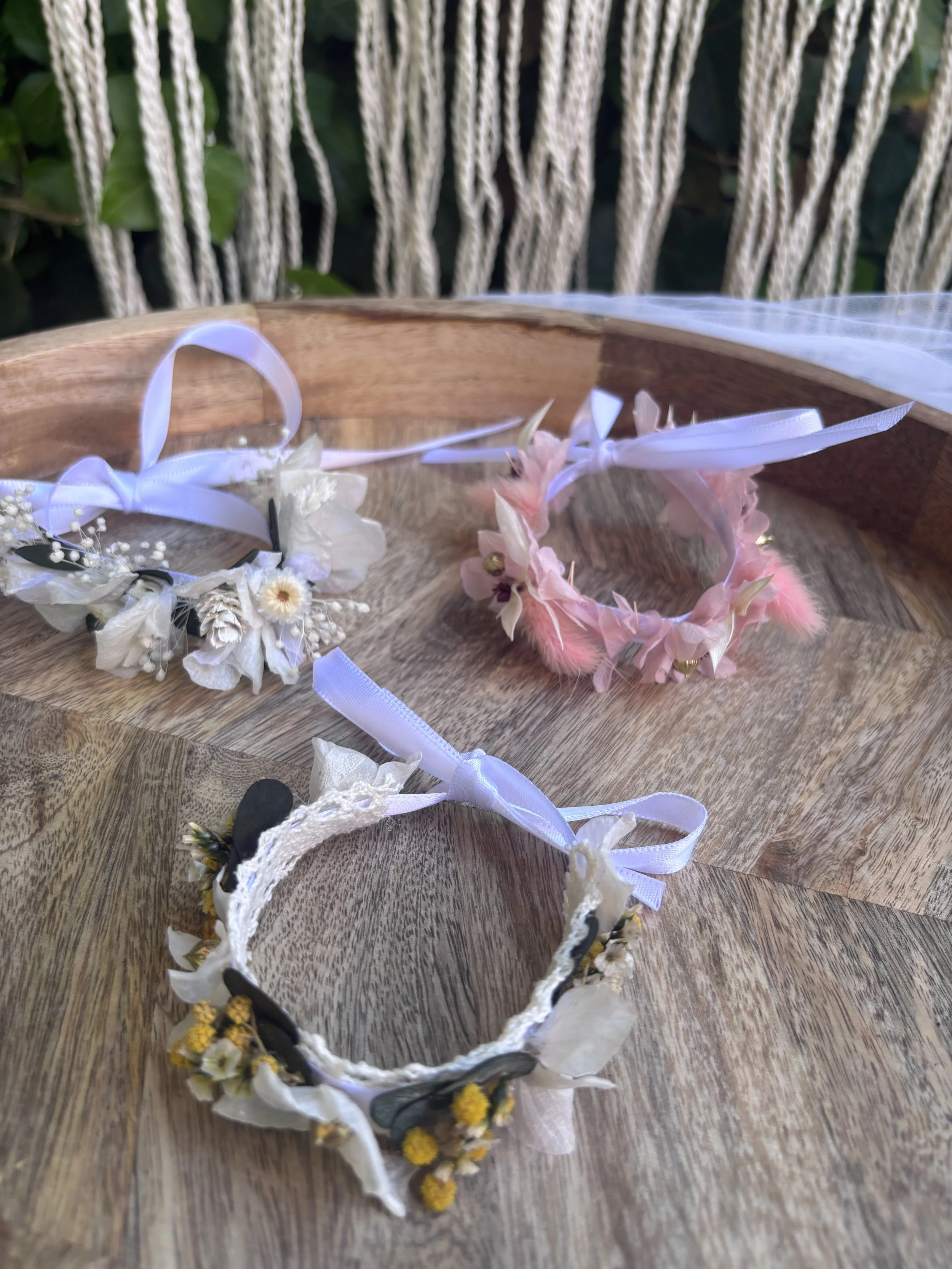 Diadèmes de fleurs et de rubans posés sur une table en bois, avec un fond de clôture en corde et de la verdure.