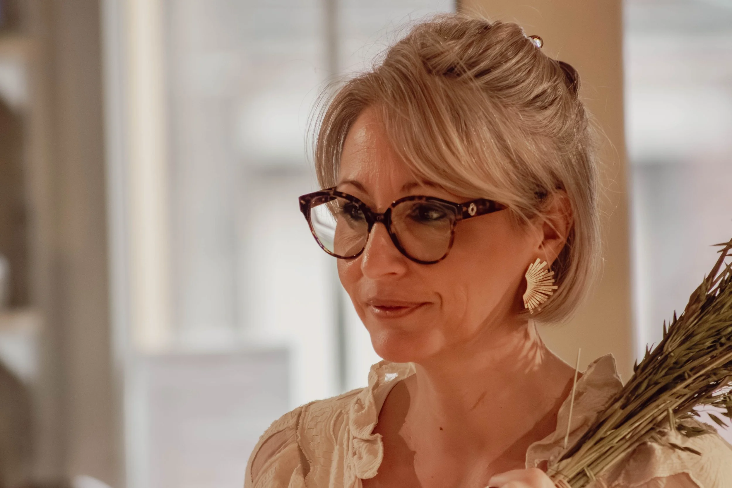 Une femme avec des cheveux blonds, portant des lunettes en écaille, sourit légèrement, portant une robe beige avec des détails en tissu.