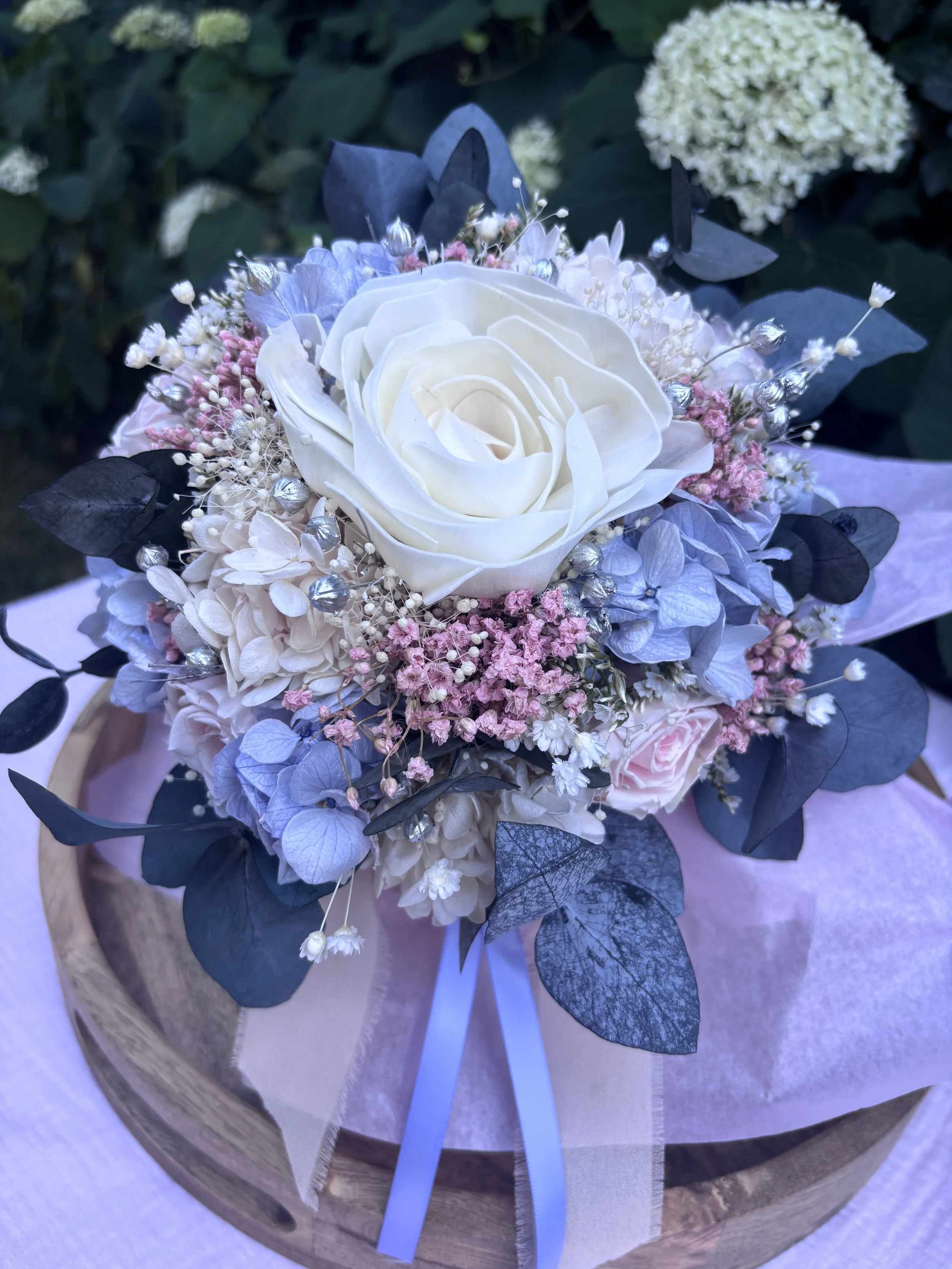 Bouquet de mariée en fleurs séchées et stabilisées aux teintes bleu pastel, rose poudré et ivoire, composé d’un camélia blanc en bois de Sola, d’hortensias stabilisés, de gypsophile, d’eucalyptus bleuté et de touches argentées. Une création romantiqu