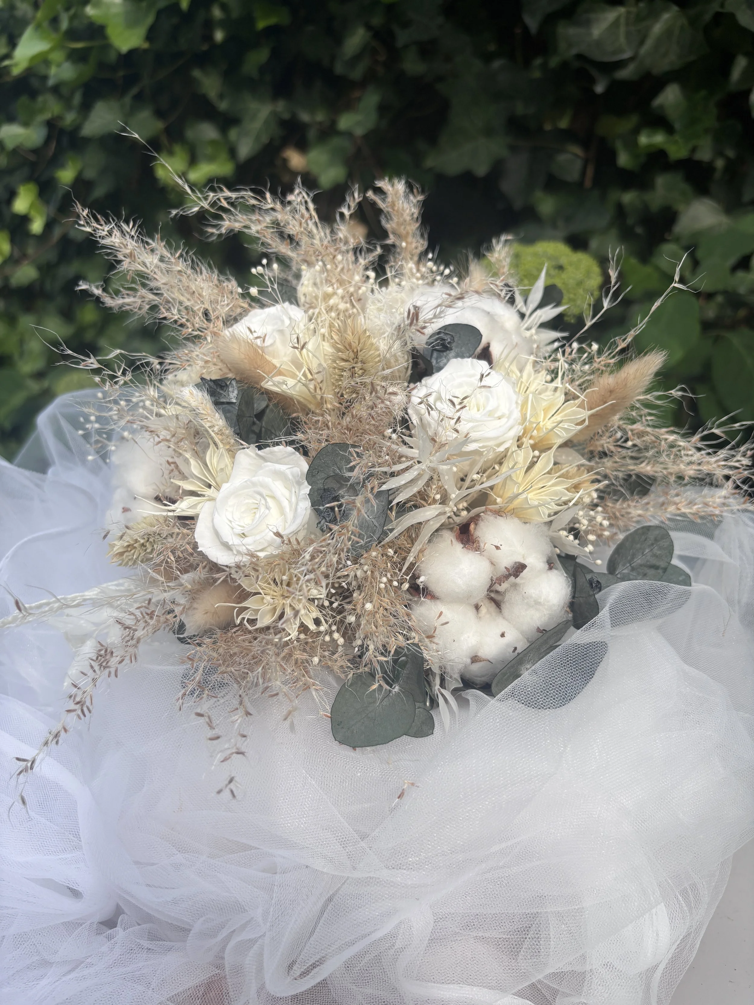 Bouquet de mariée bohème chic réalisé en fleurs séchées et stabilisées dans des nuances de blanc, ivoire, beige naturel et vert eucalyptus. Cette création artisanale associe roses stabilisées, coton, graminées aériennes, eucalyptus et fleurs texturée