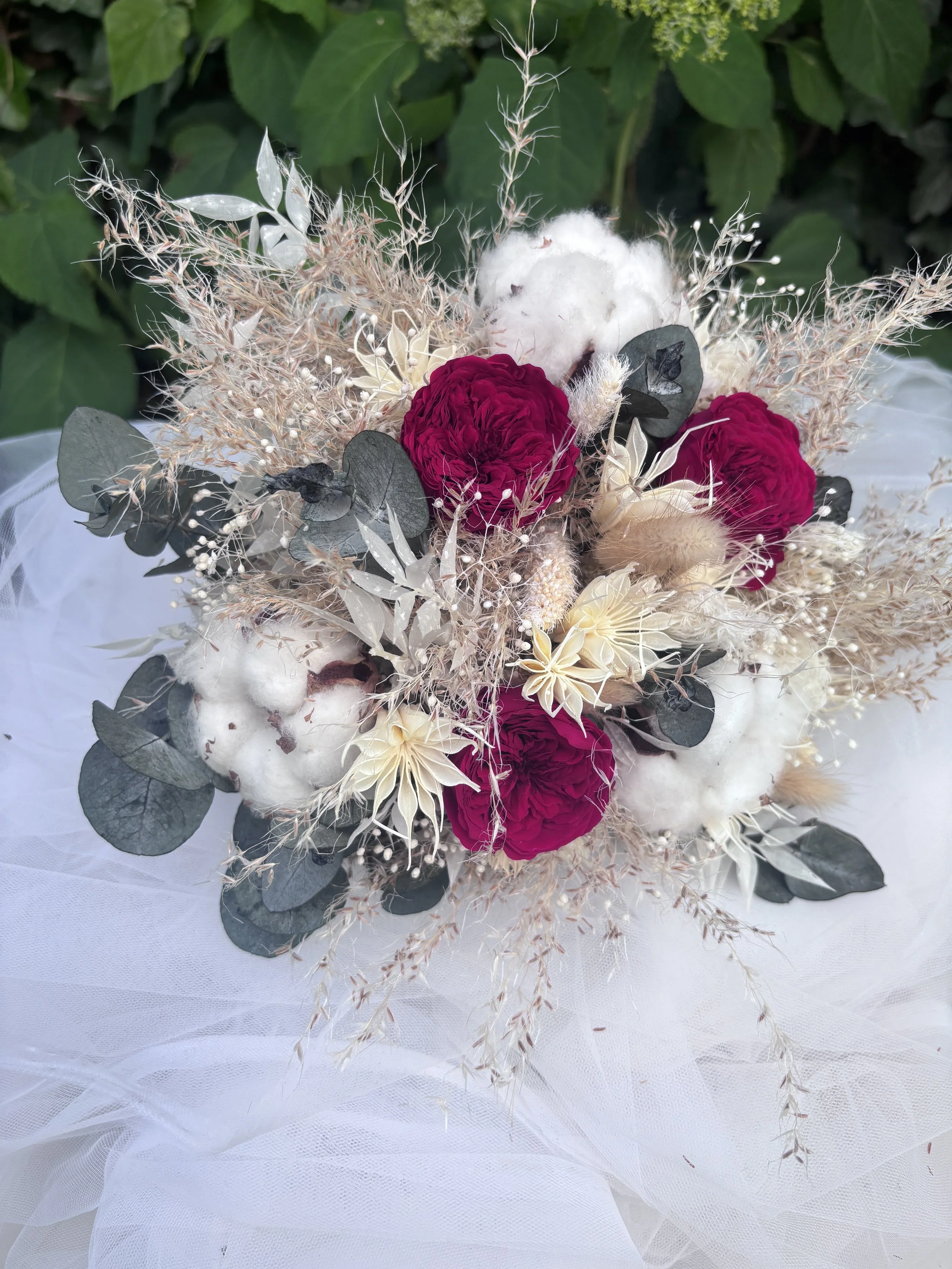 Bouquet de mariée bohème aux nuances bordeaux profond, ivoire et beige naturel, réalisé à partir de fleurs séchées et stabilisées. Cette création florale associe roses stabilisées bordeaux, coton, eucalyptus, graminées aériennes et fleurs texturées p