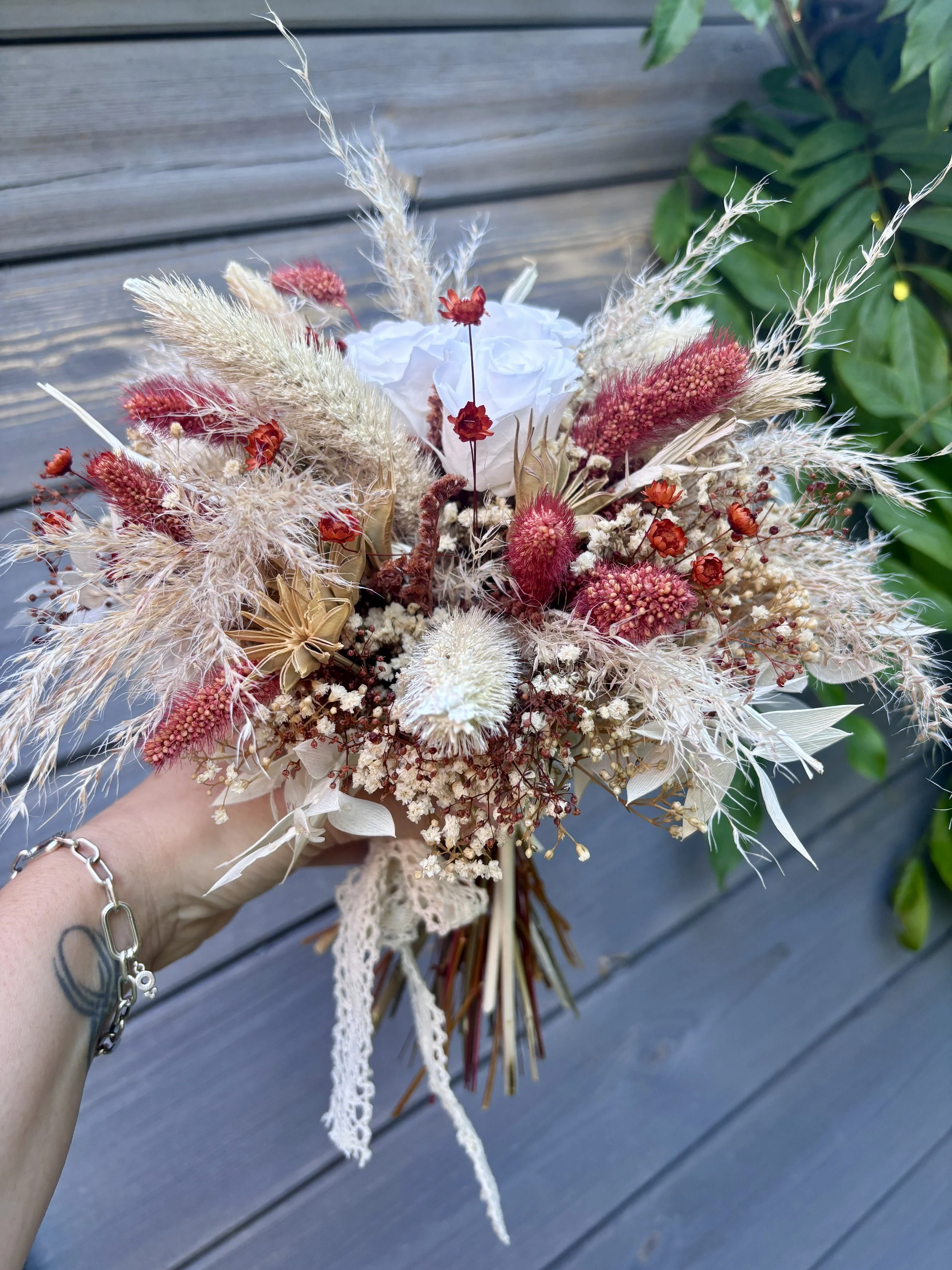 Bouquet de mariée bohème en fleurs séchées et stabilisées aux teintes terracotta, bordeaux, crème et blanc. Cette création florale artisanale associe roses éternelles blanches, pampas, setaria, graminées naturelles et délicates fleurs séchées pour of