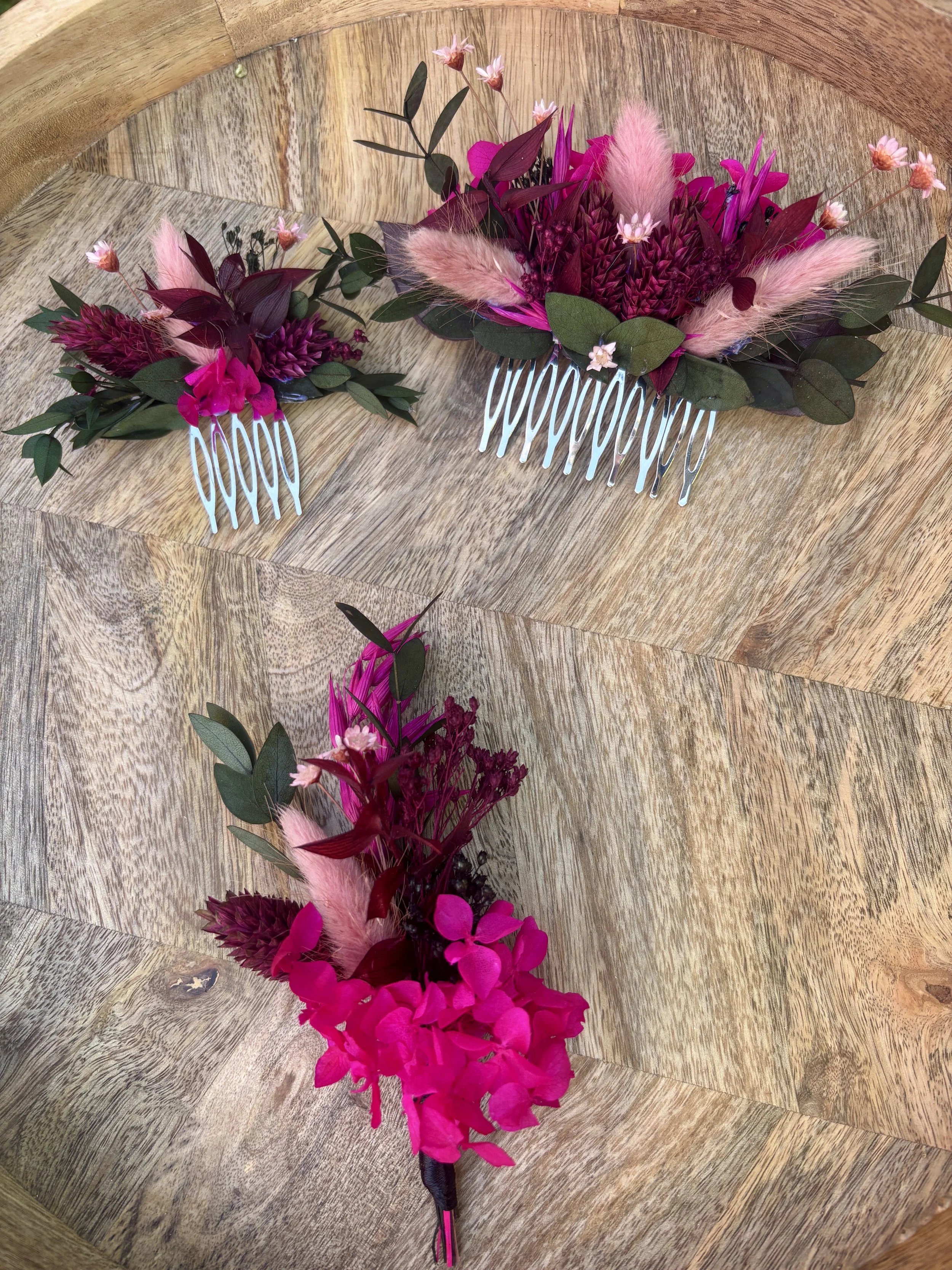 Un assortiment de fleurs roses, fushia, et vertes avec des feuilles, arrangé dans deux peignes à cheveux, posé sur une surface en bois.