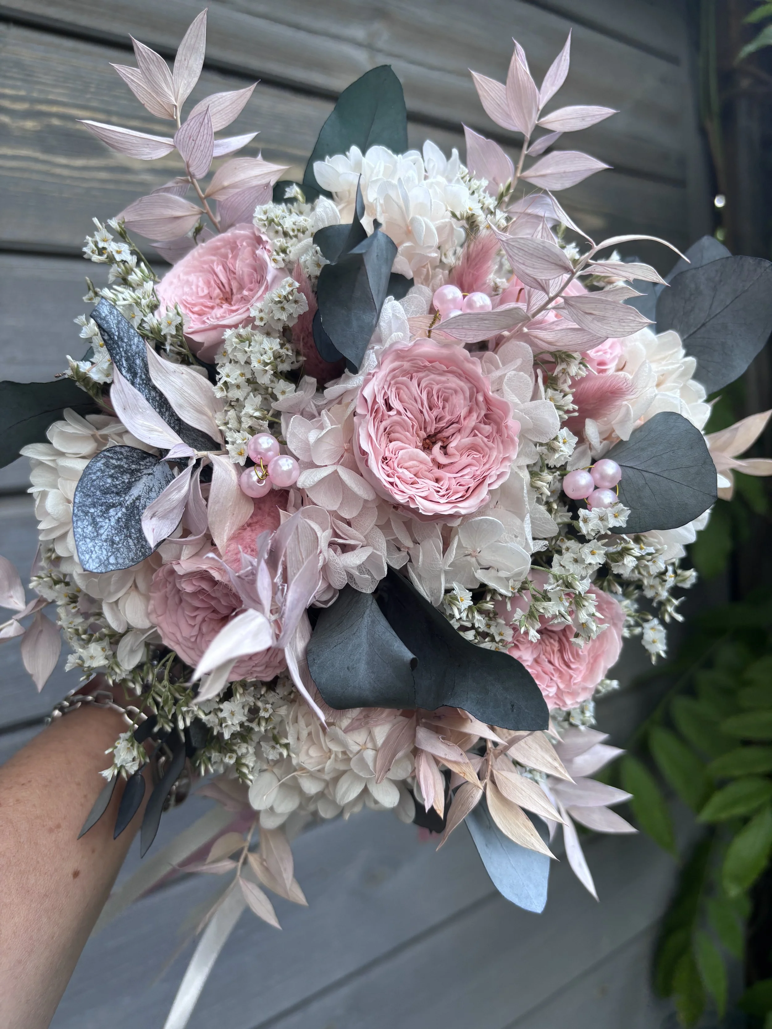 Bouquet de mariée bohème réalisé à la main en fleurs séchées et stabilisées dans des tons rose poudré, blanc et gris bleuté. Cette création florale associe de délicates roses anglaises stabilisées, des hortensias préservés, de l’eucalyptus et des feu