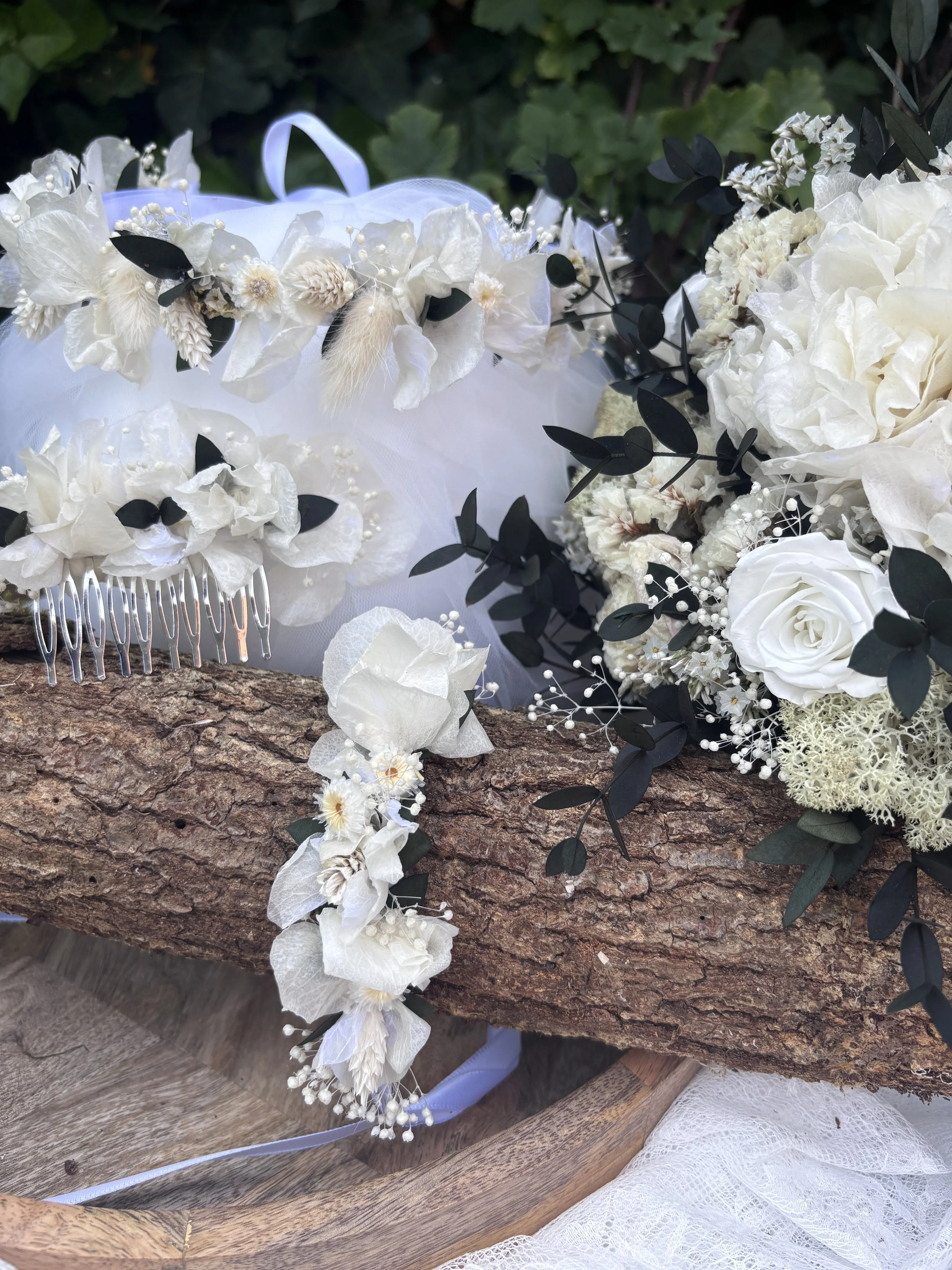 Un bouquet de fleurs blanches avec des feuilles vertes, posé sur du bois, accompagnée d'une pièce de tissu en dentelle et d'une pince à cheveux en métal.