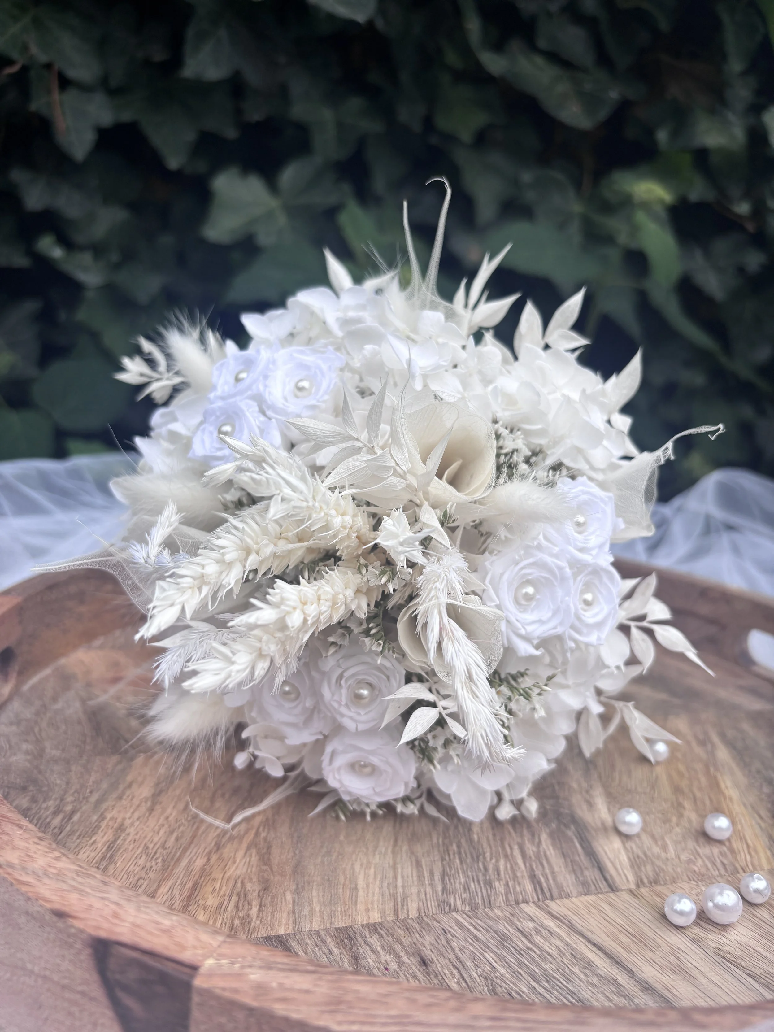 Bouquet de mariée en fleurs séchées et stabilisées blanches, réalisé artisanalement dans un esprit bohème chic et romantique. Cette création florale associe hortensias stabilisés, roses éternelles ornées de perles, arums délicats, feuillages blanchis