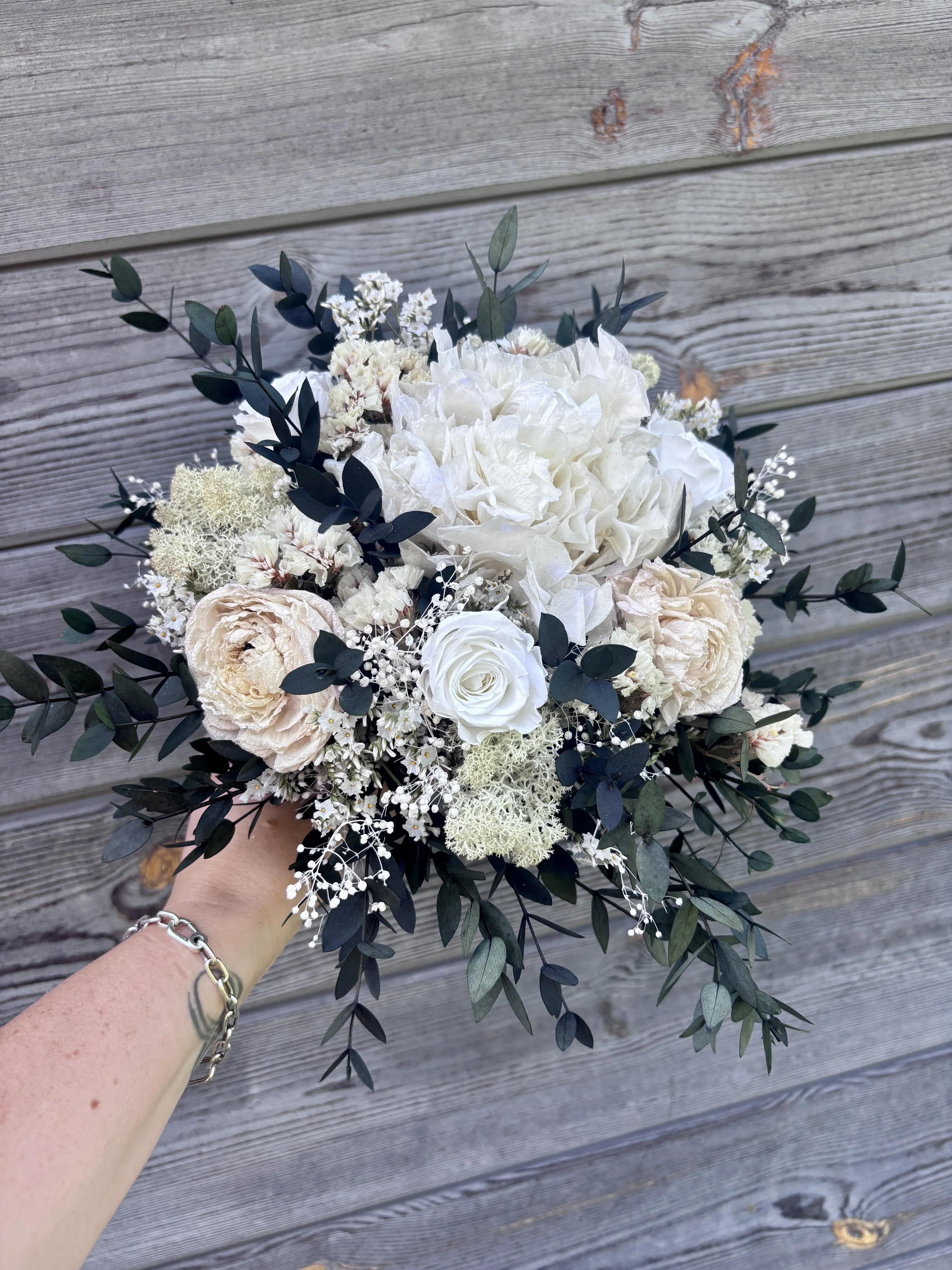 Bouquet de mariée bohème chic en fleurs séchées et stabilisées, composé de pivoines blanches, roses stabilisées, statice blanc, mousse végétale et feuillage d’eucalyptus noir. Cette création artisanale associe élégance, douceur et caractère pour un m