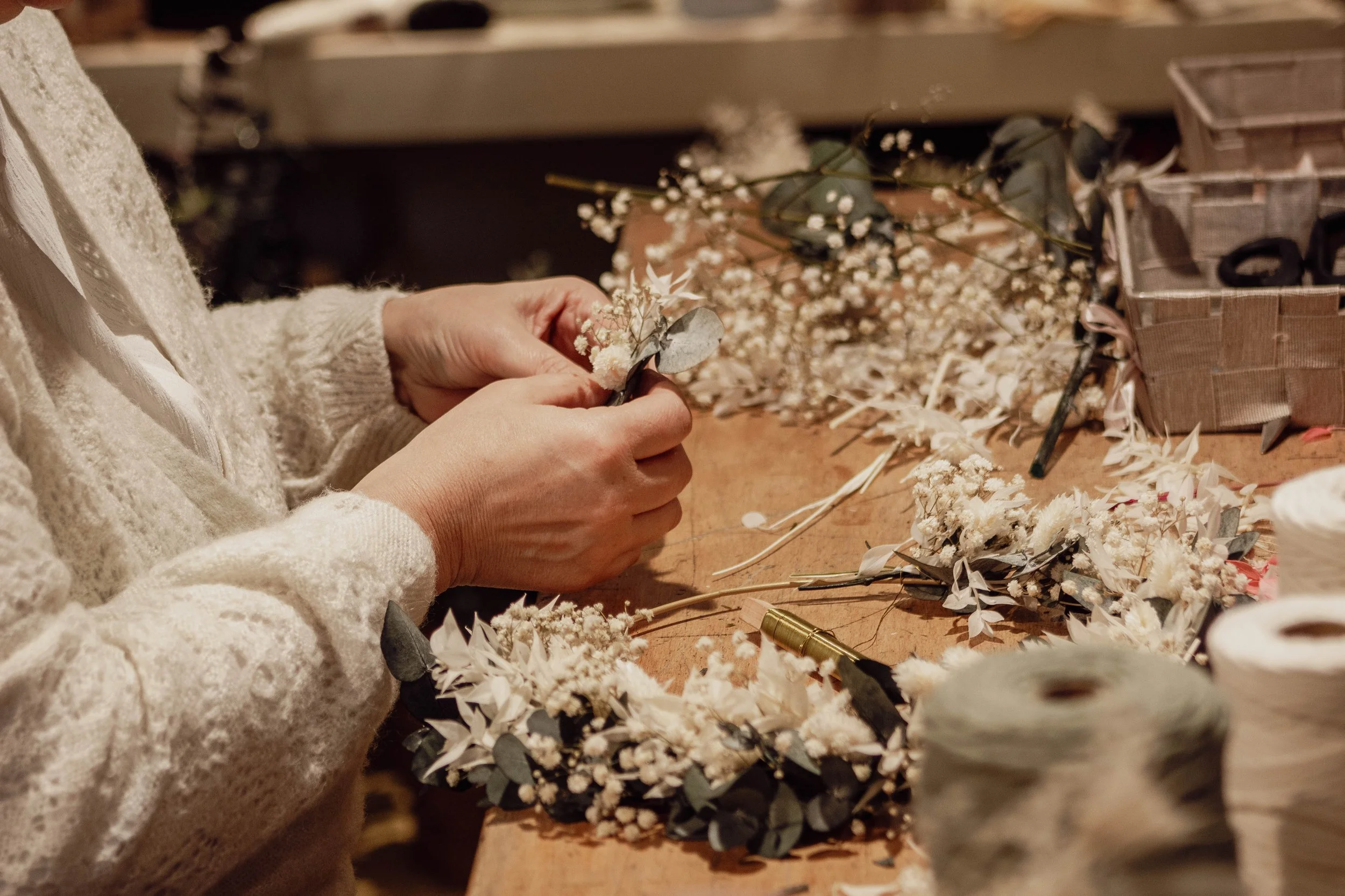 Personne faisant un arrangement floral avec des fleurs blanches et des feuilles. Plan rapproché des mains, avec de nombreux outils et fleurs éparpillés sur une table en bois.
