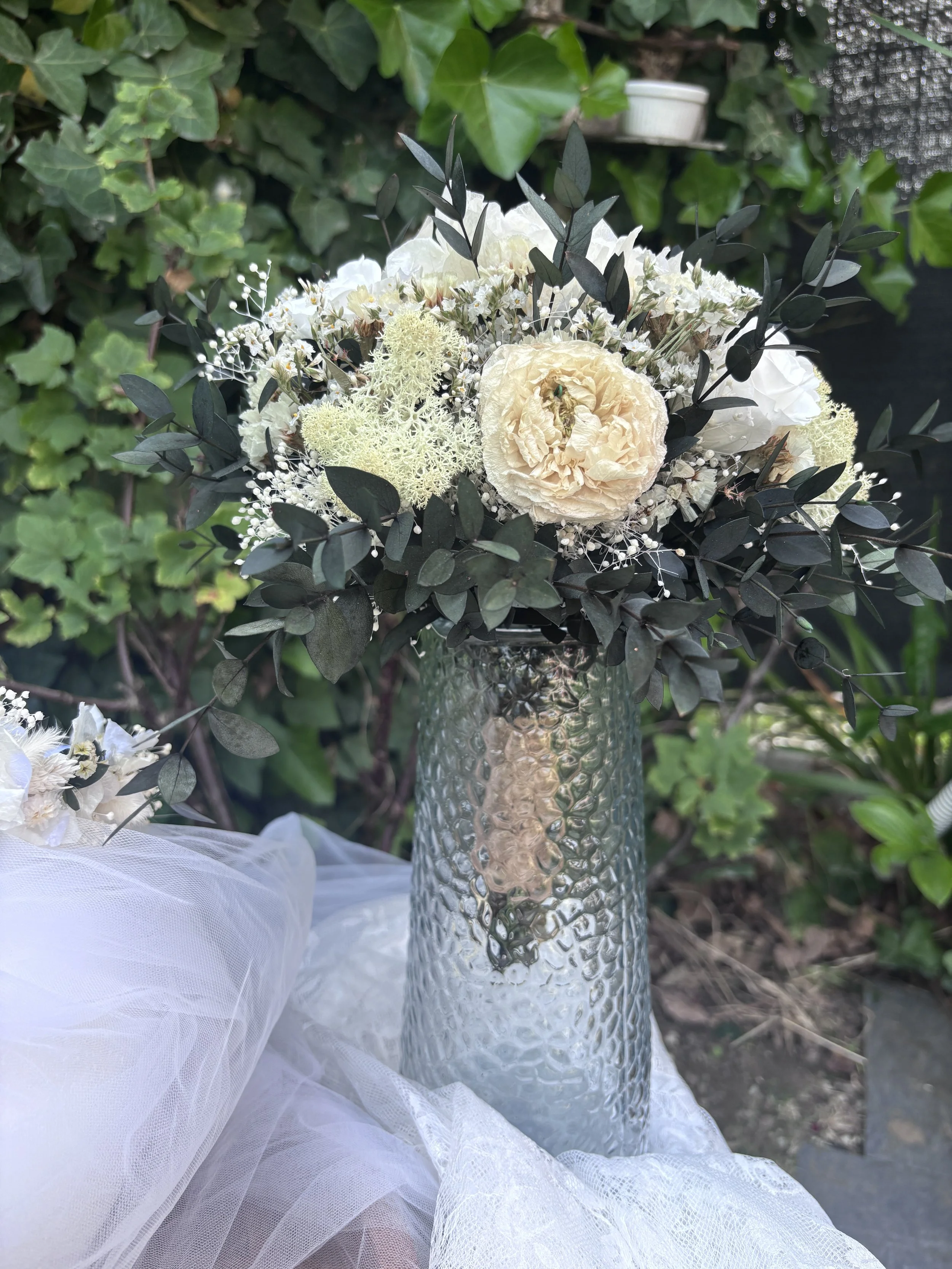 Bouquet de mariée bohème chic en fleurs séchées et stabilisées, composé de pivoines blanches, roses stabilisées, statice blanc, mousse végétale et feuillage d’eucalyptus noir. Cette création artisanale associe élégance, douceur et caractère pour un m