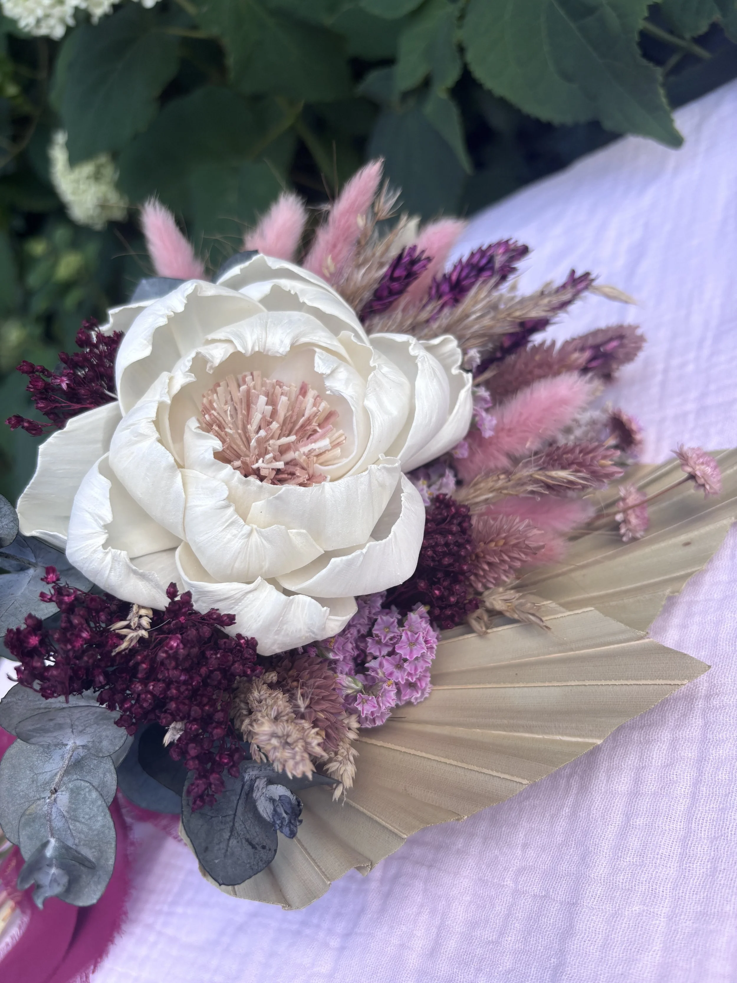 Bouquet de mariée en fleurs séchées et stabilisées, composé d’une grande pivoine ivoire en bois de Sola, d’eucalyptus, de lagurus rose poudré, de fleurs bordeaux et de palmes naturelles. Un bouquet romantique et durable, idéal pour un mariage bohème 