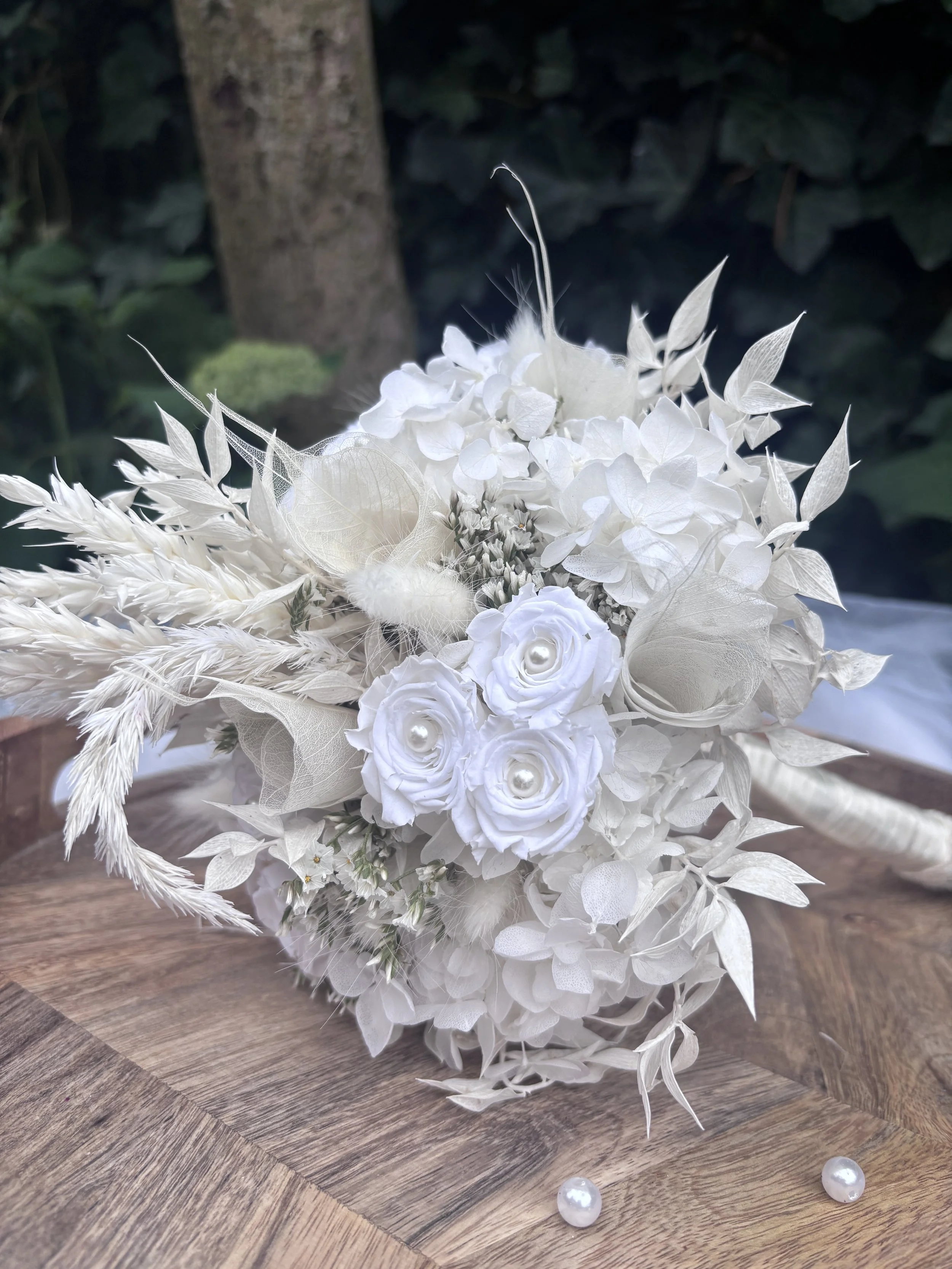 Bouquet de mariée en fleurs séchées et stabilisées blanches, réalisé artisanalement dans un esprit bohème chic et romantique. Cette création florale associe hortensias stabilisés, roses éternelles ornées de perles, arums délicats, feuillages blanchis