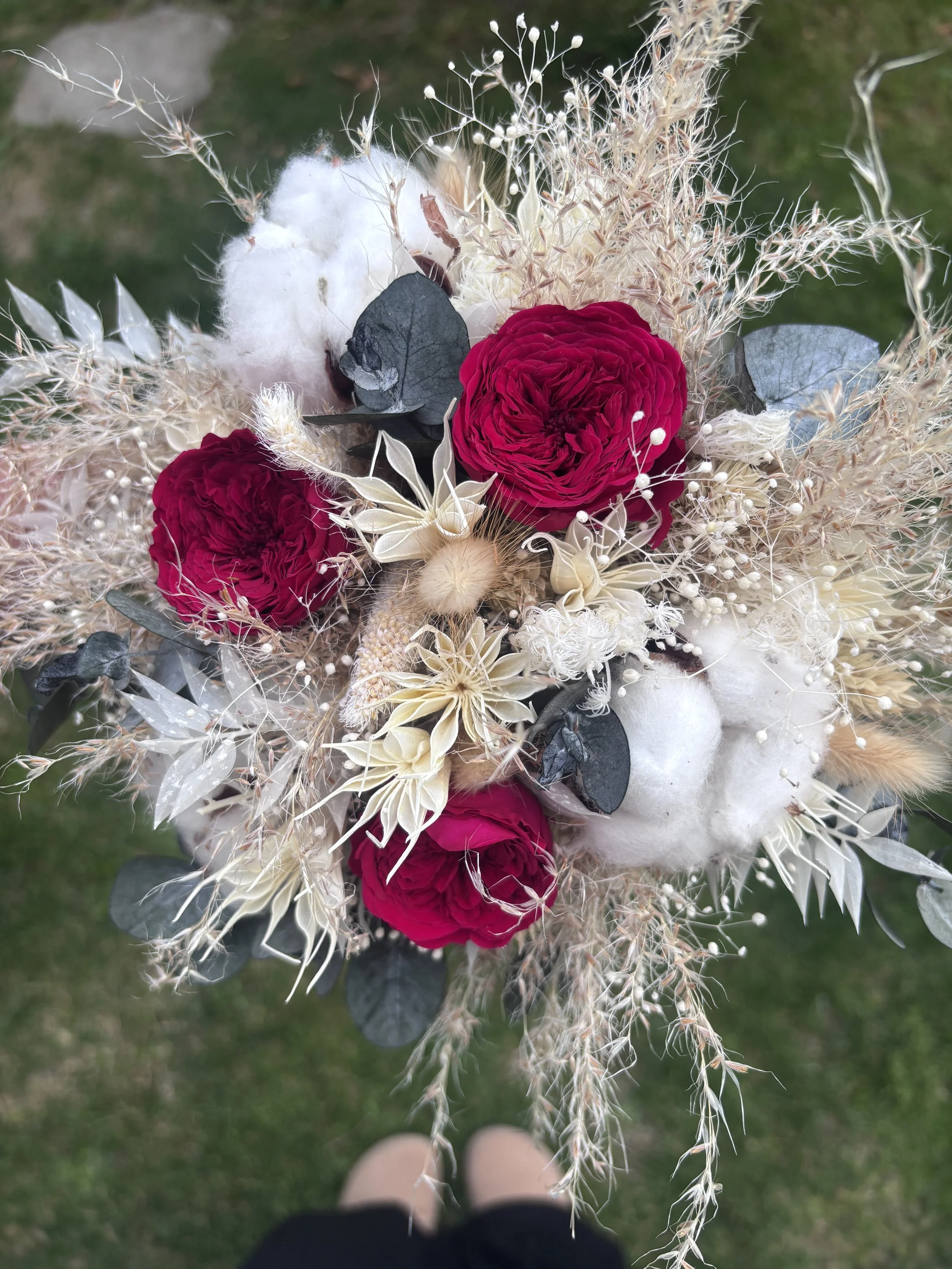 Bouquet de mariée bohème aux nuances bordeaux profond, ivoire et beige naturel, réalisé à partir de fleurs séchées et stabilisées. Cette création florale associe roses stabilisées bordeaux, coton, eucalyptus, graminées aériennes et fleurs texturées p