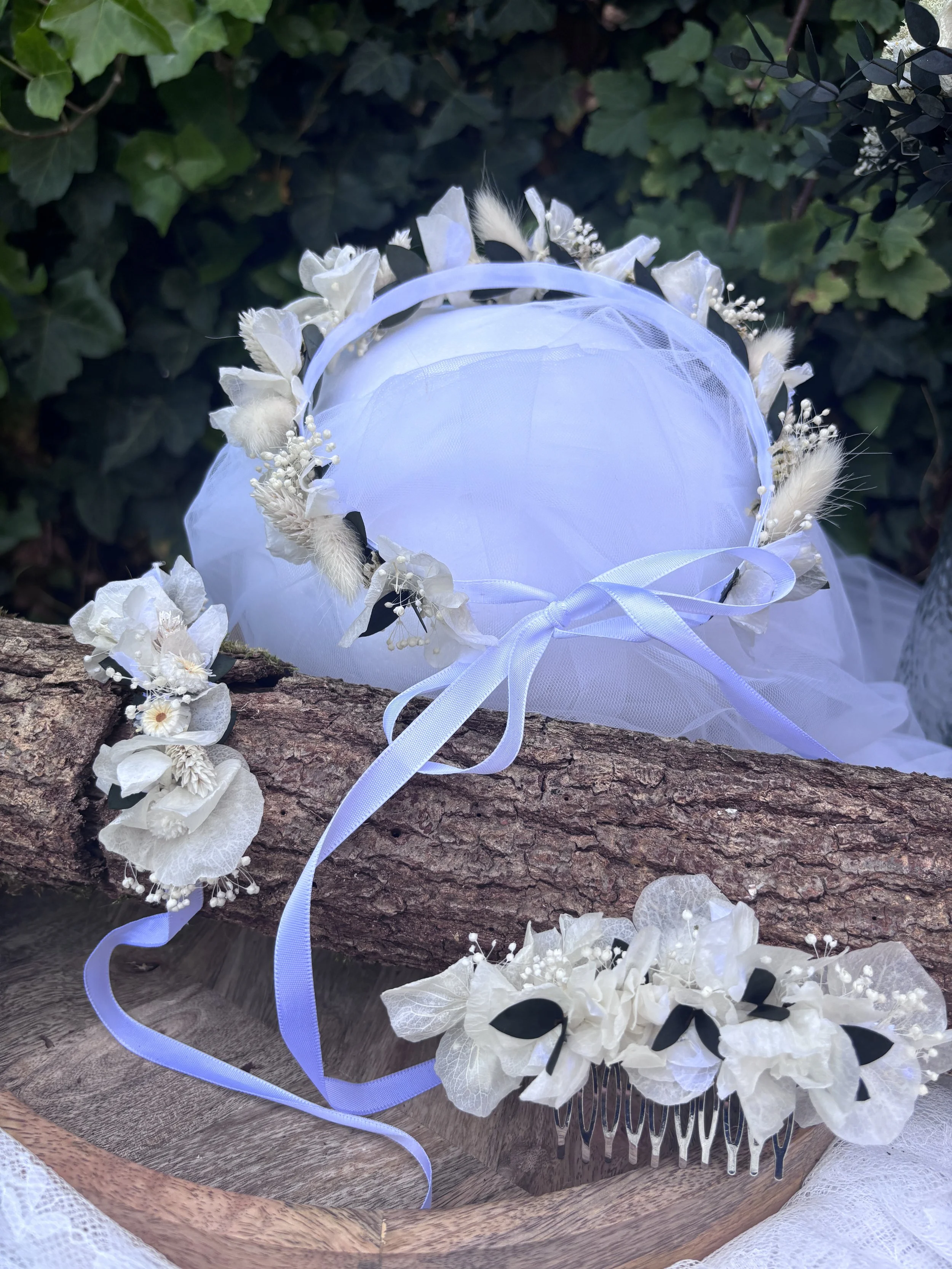 Un diadème blanc avec des fleurs et des feuilles, posé sur une branche en bois, avec un fond de feuillage vert.