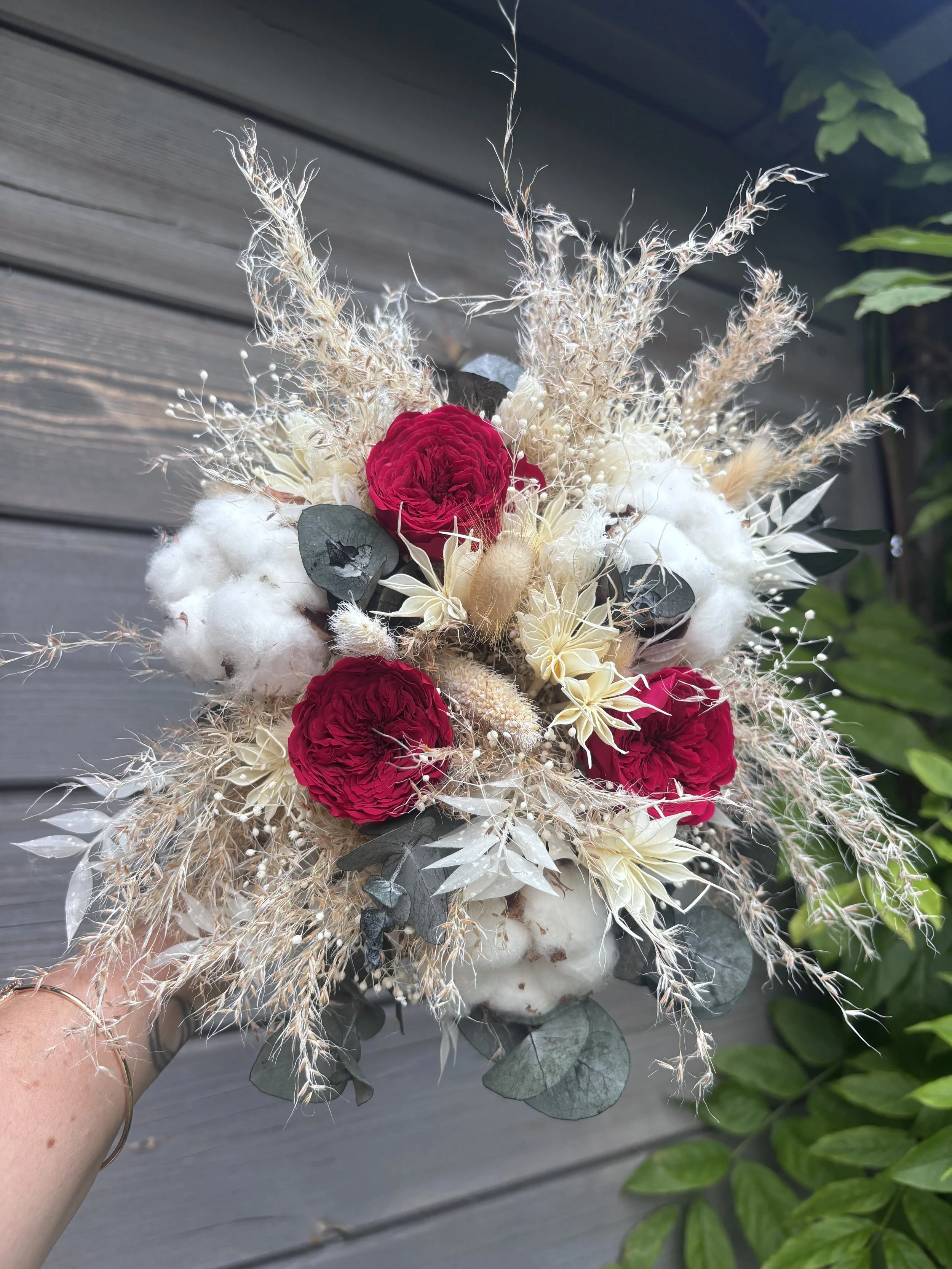 Bouquet de mariée bohème aux nuances bordeaux profond, ivoire et beige naturel, réalisé à partir de fleurs séchées et stabilisées. Cette création florale associe roses stabilisées bordeaux, coton, eucalyptus, graminées aériennes et fleurs texturées p