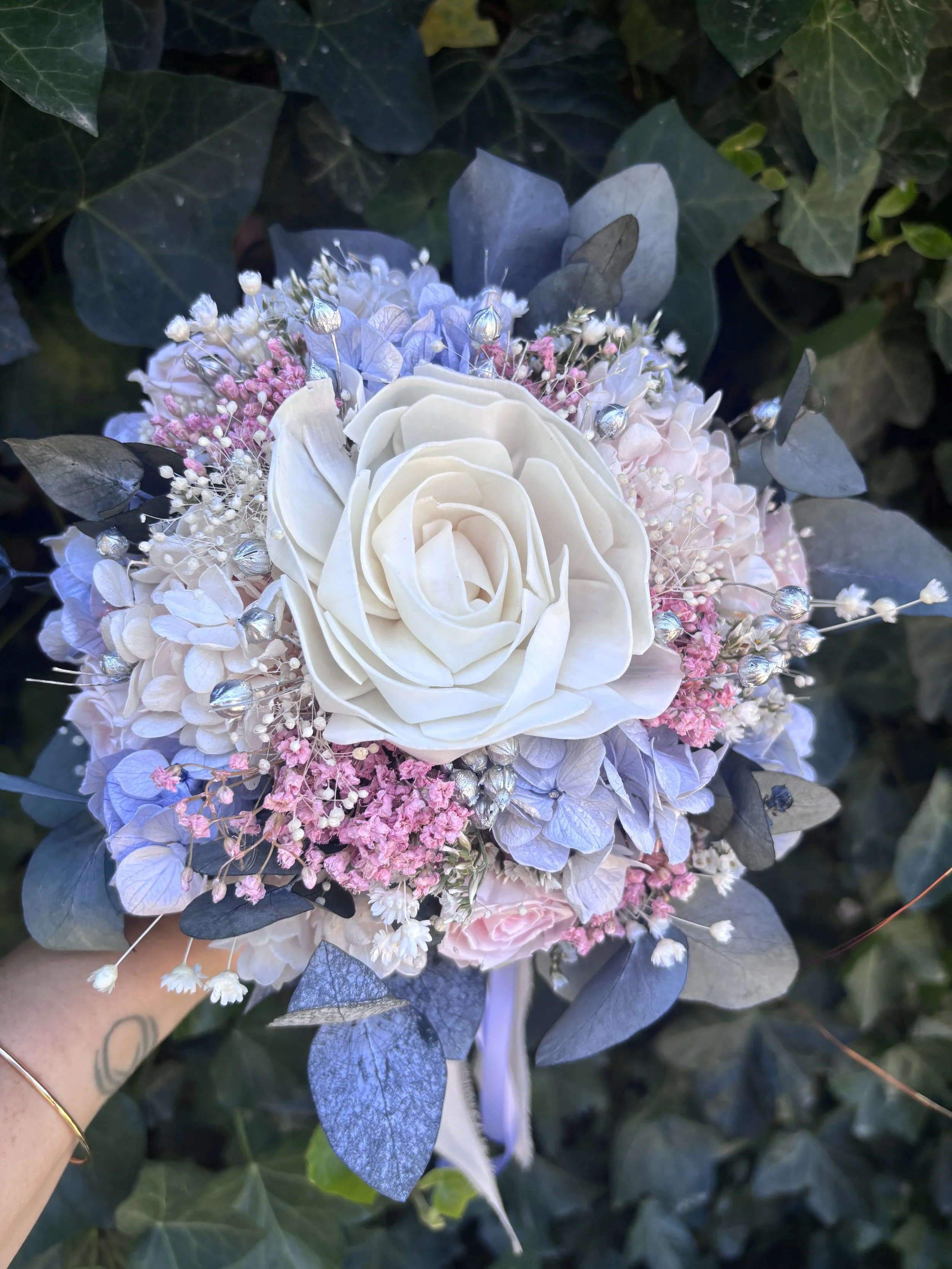 Bouquet de mariée en fleurs séchées et stabilisées aux teintes bleu pastel, rose poudré et ivoire, composé d’un camélia blanc en bois de Sola, d’hortensias stabilisés, de gypsophile, d’eucalyptus bleuté et de touches argentées. Une création romantiqu