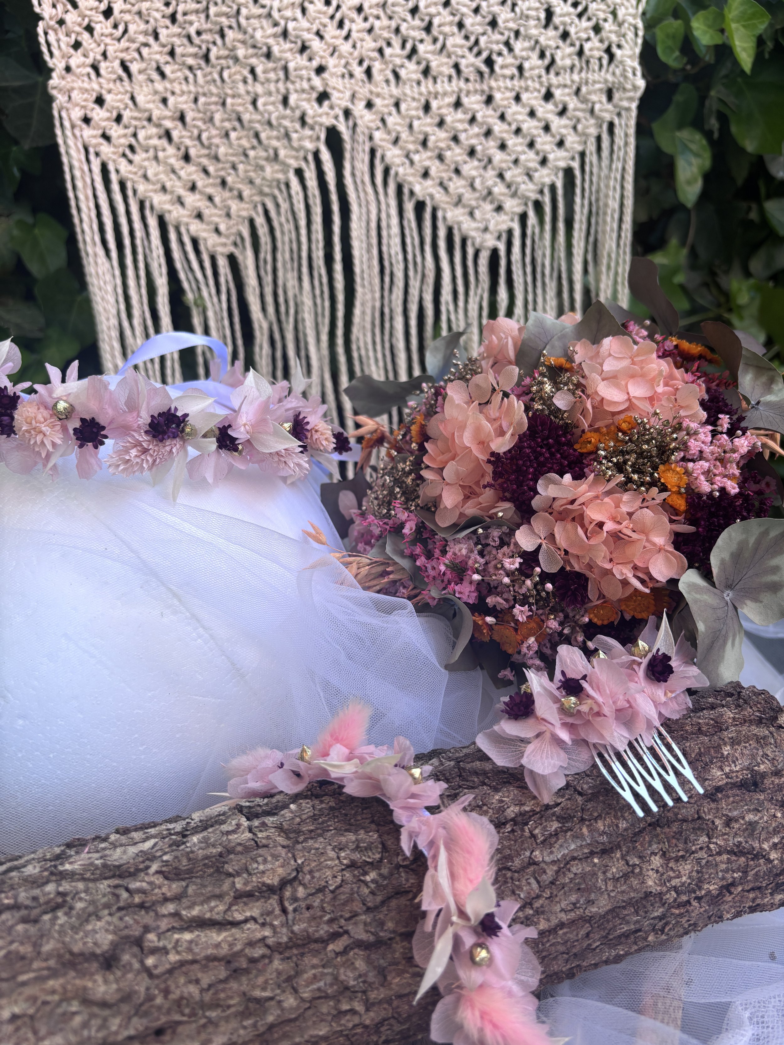 Couronne de fleurs roses, violettes et orange posée sur une branche de bois, avec un fond de tissu blanc et un panier en macramé beige.