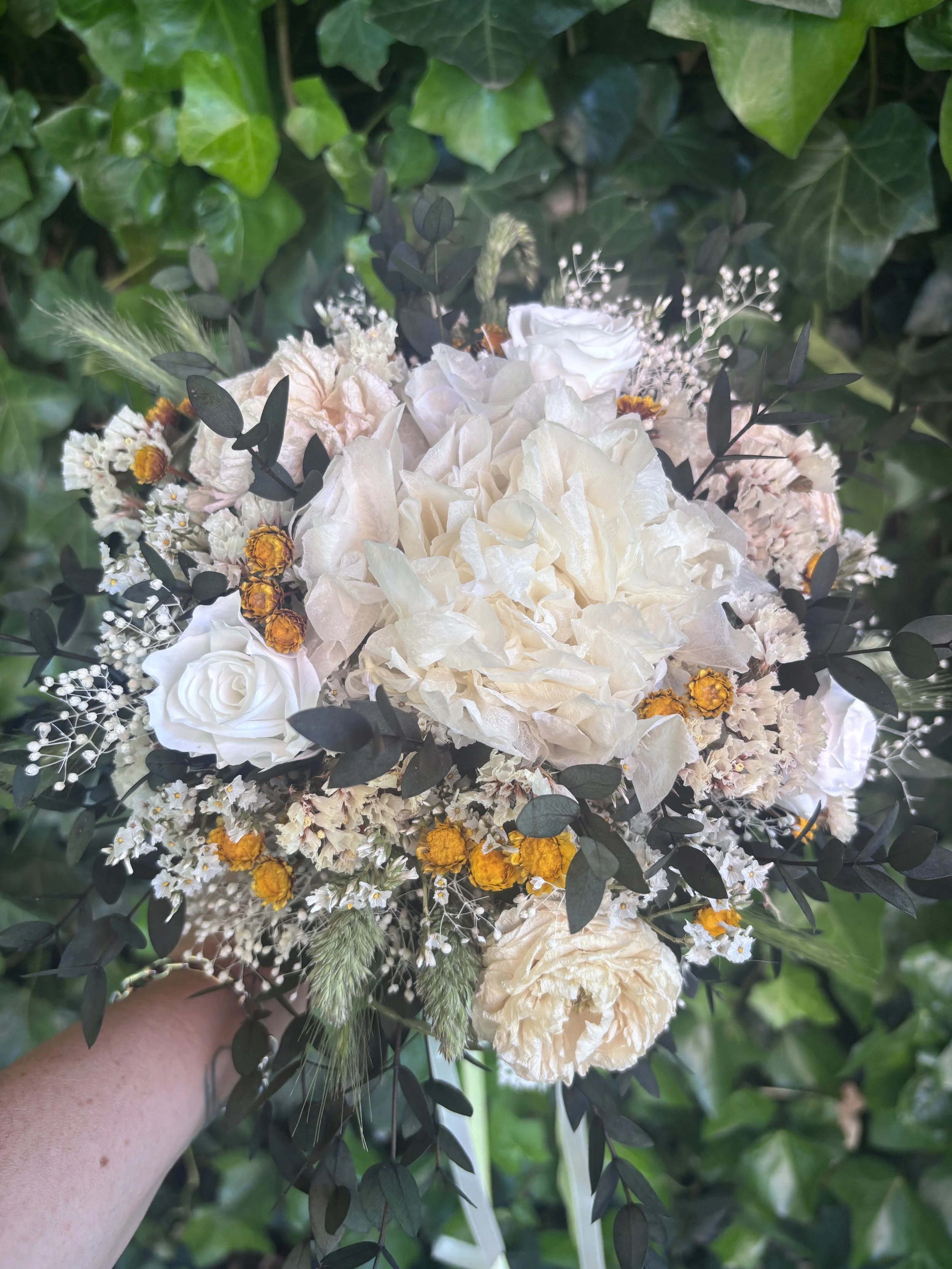 Bouquet de mariée bohème en fleurs séchées et stabilisées, réalisé à la main dans des tons ivoire, blanc cassé, crème et jaune doré. Cette création florale associe hortensia stabilisé, roses éternelles blanches, renoncules séchées, statice, graminées