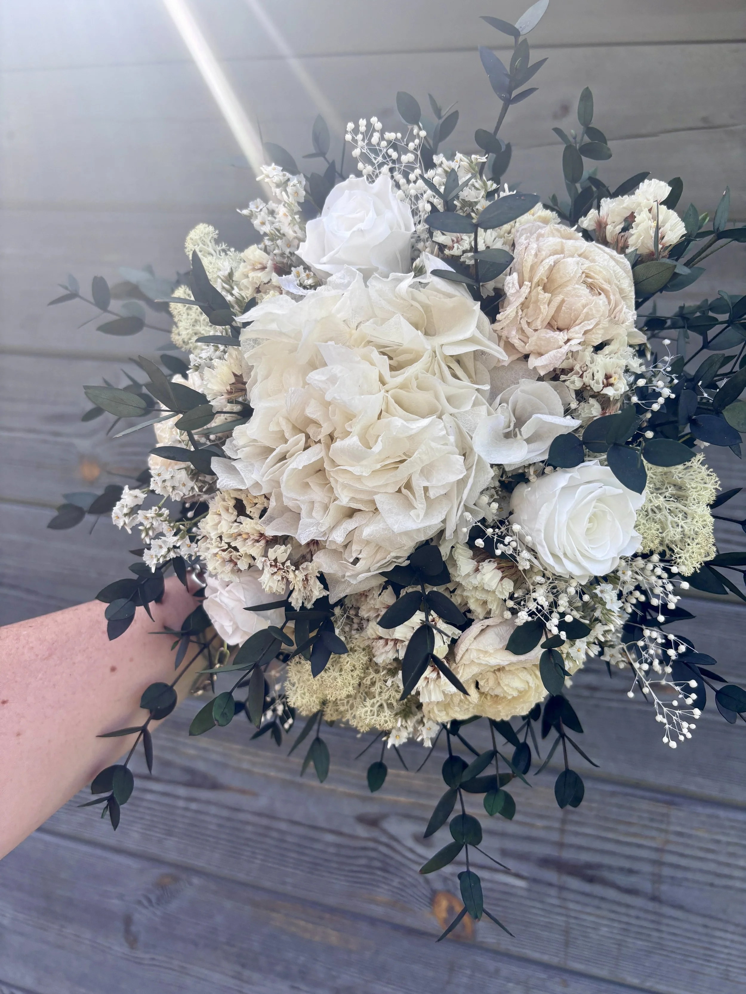 Bouquet de mariée bohème chic en fleurs séchées et stabilisées, composé de pivoines blanches, roses stabilisées, statice blanc, mousse végétale et feuillage d’eucalyptus noir. Cette création artisanale associe élégance, douceur et caractère pour un m