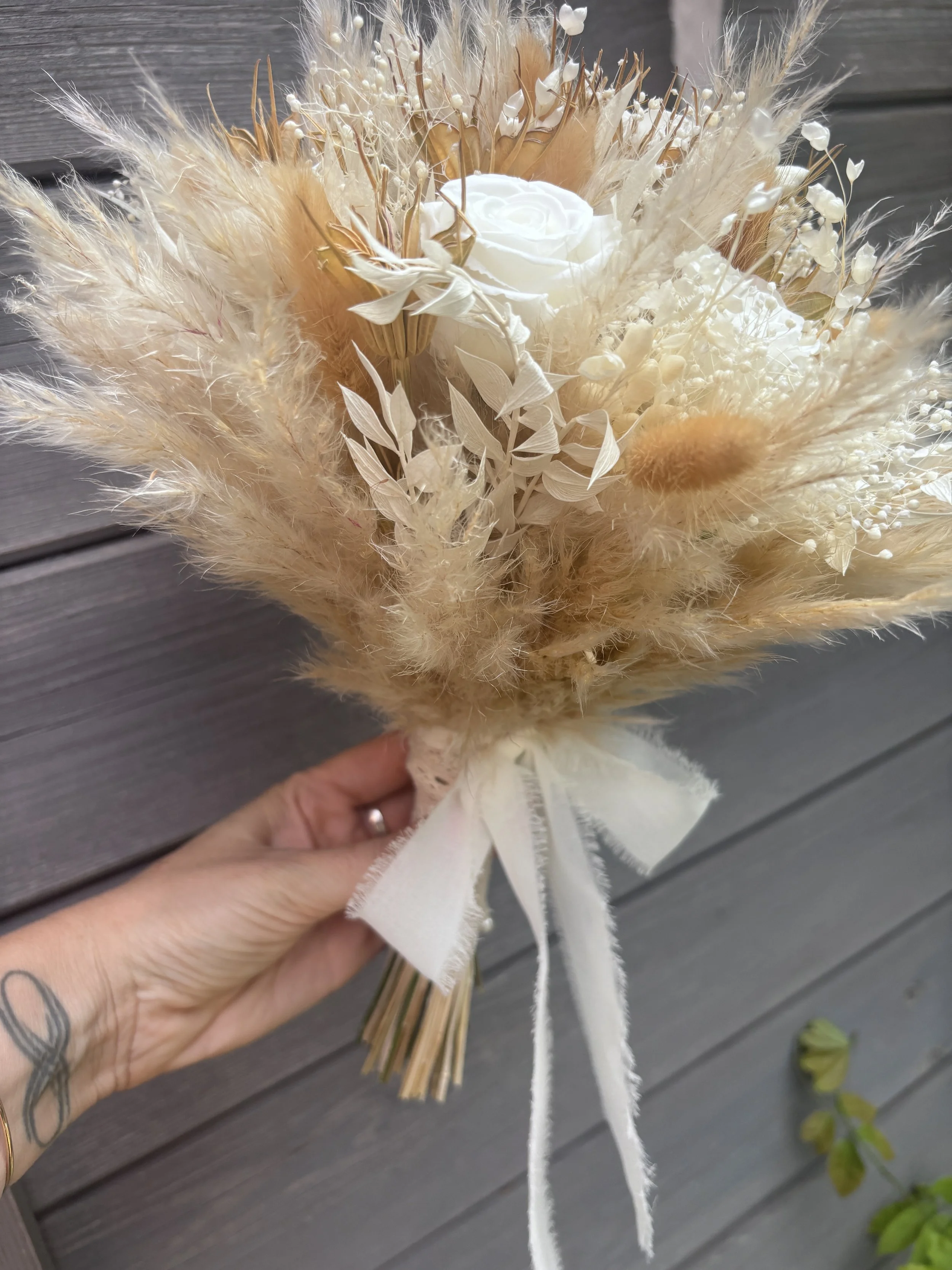 Bouquet de mariée réalisé artisanalement en fleurs séchées et stabilisées. Une création romantique et aérienne mêlant roses blanches, gypsophile, pampas et feuillages naturels dans des nuances ivoire, nude et rose poudré. Création sur mesure par L’At