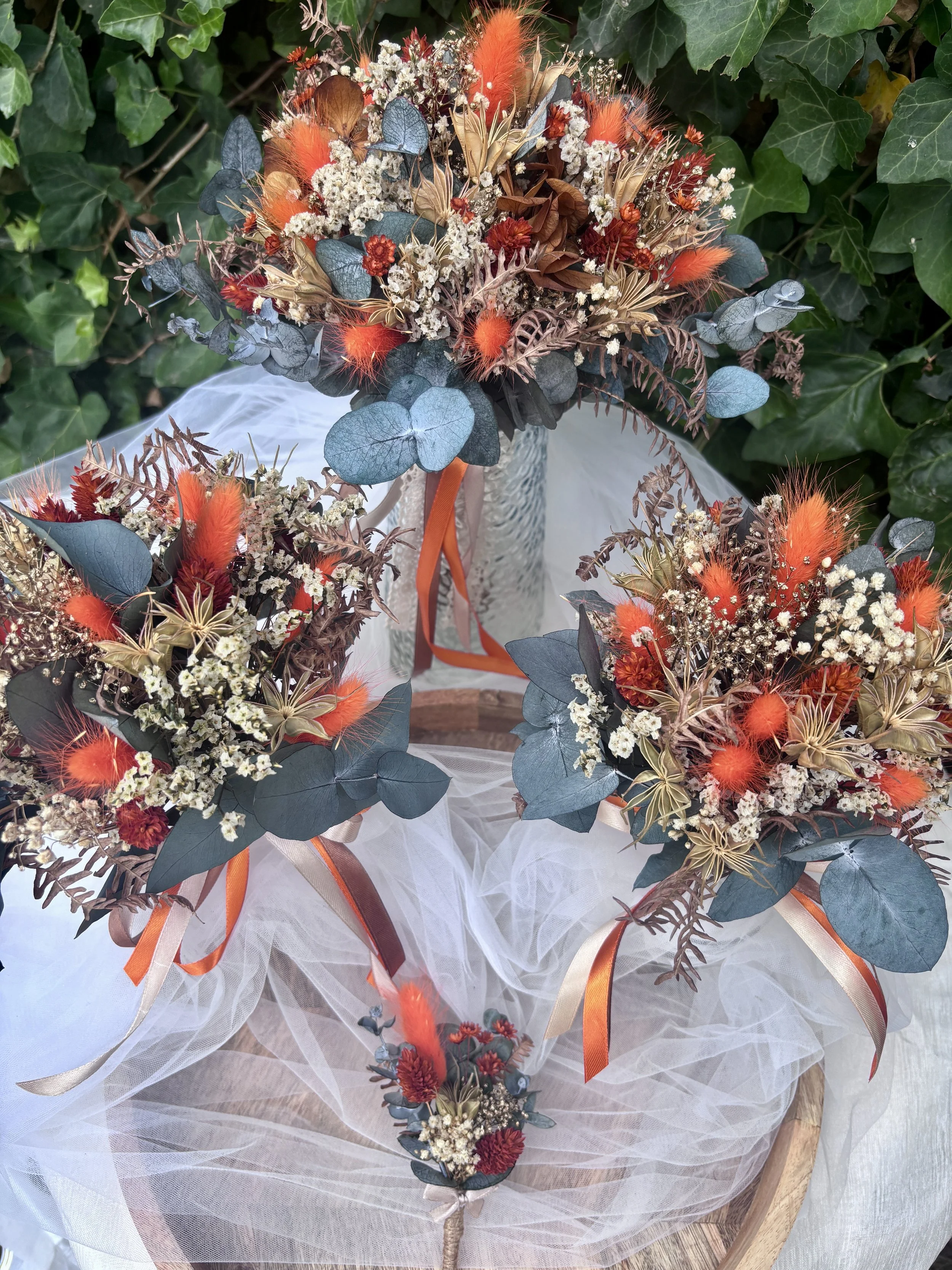 Ensemble floral coordonné composé d’un bouquet de mariée, de deux bouquets de demoiselles d’honneur et d’une boutonnière pour le marié. Réalisées artisanalement en fleurs séchées et stabilisées, ces créations associent des nuances terracotta, orange,