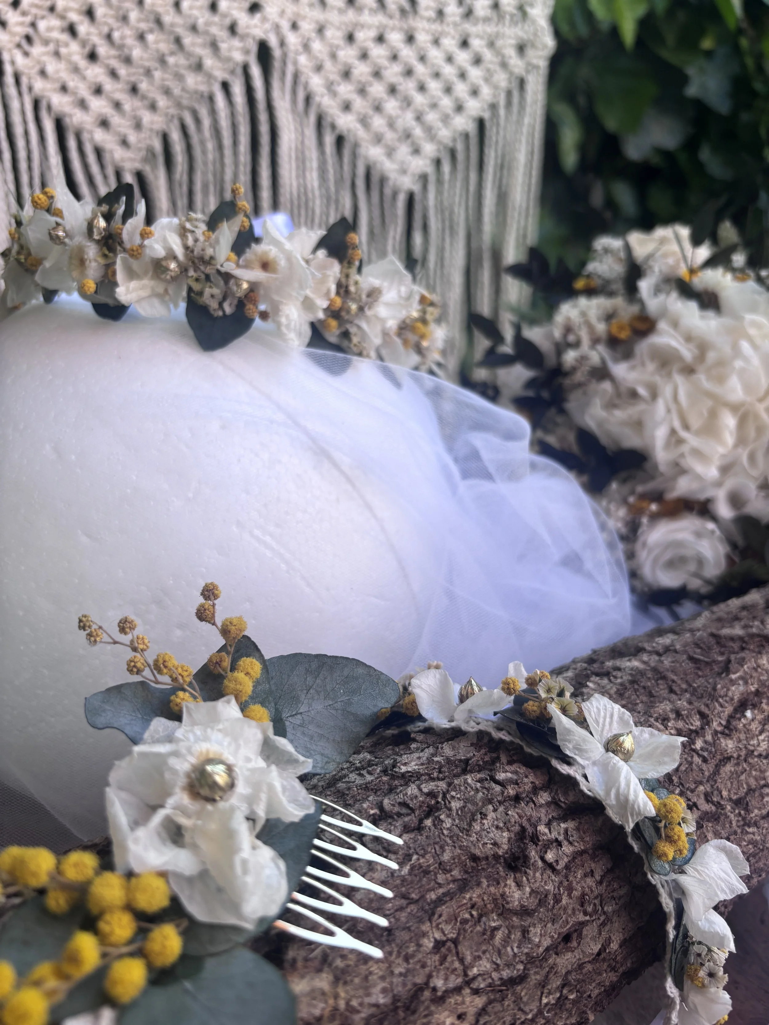 Une couronne de fleurs blanches et jaunes posée sur une table en bois, avec un fond flou de plantes vertes.