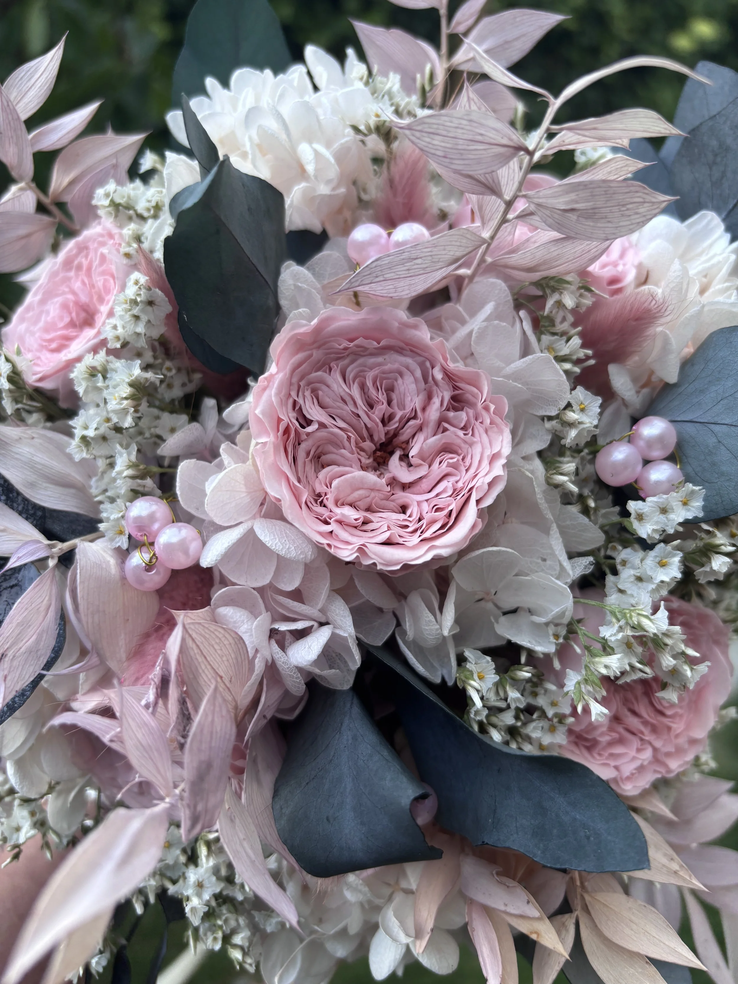 Bouquet de mariée bohème réalisé à la main en fleurs séchées et stabilisées dans des tons rose poudré, blanc et gris bleuté. Cette création florale associe de délicates roses anglaises stabilisées, des hortensias préservés, de l’eucalyptus et des feu