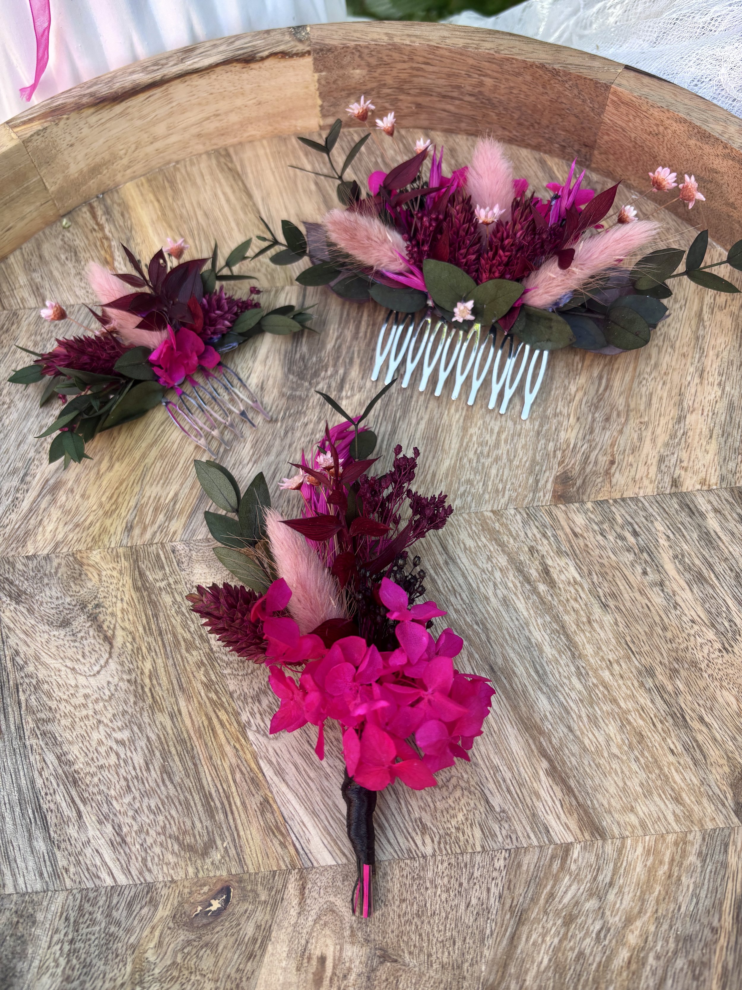 Une coiffure florale avec des fleurs roses et violettes, des feuilles vertes et des plumes roses, fixée à l'aide d'un peigne sur une surface en bois.