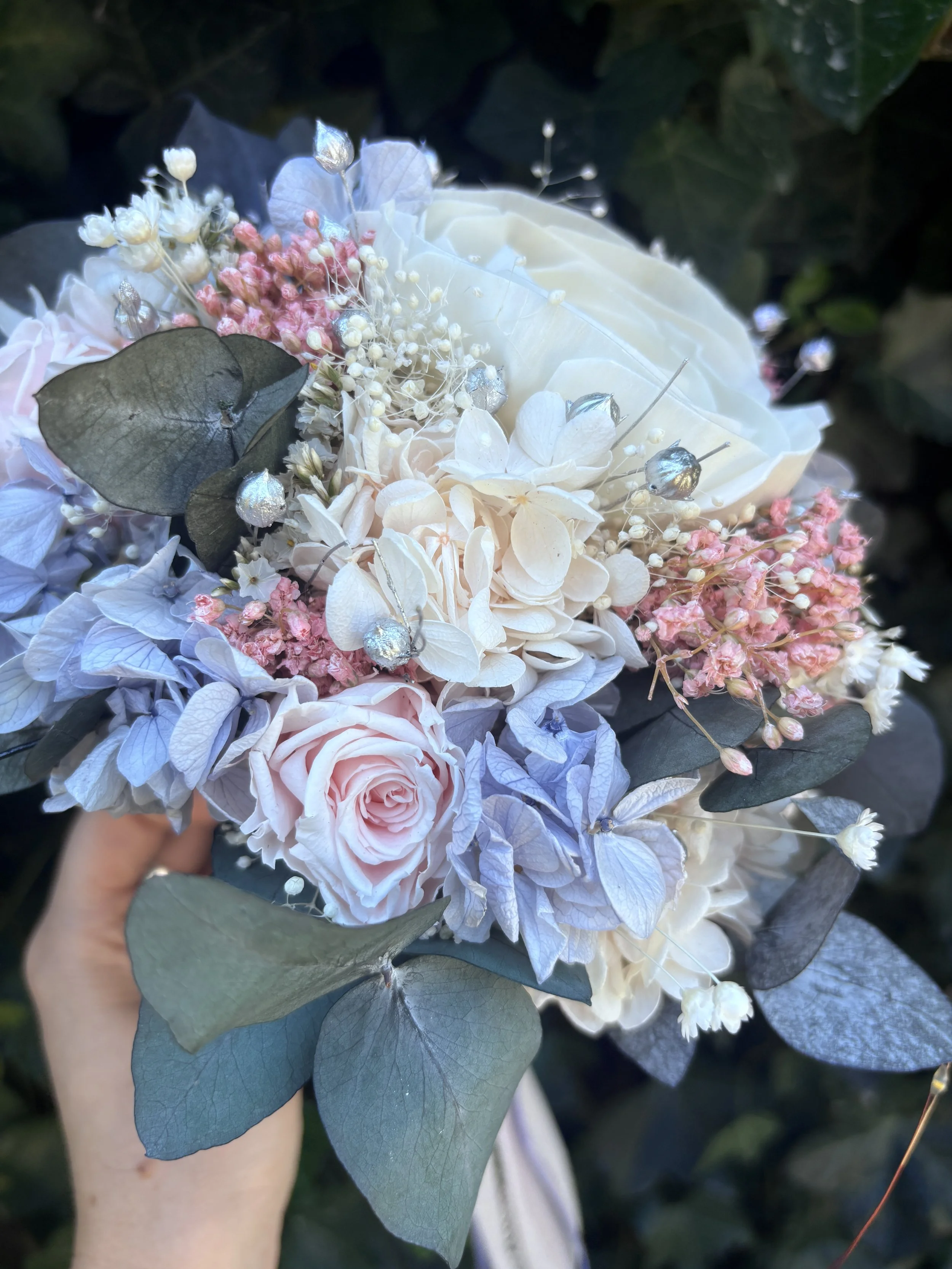 Bouquet de mariée en fleurs séchées et stabilisées aux teintes bleu pastel, rose poudré et ivoire, composé d’un camélia blanc en bois de Sola, d’hortensias stabilisés, de gypsophile, d’eucalyptus bleuté et de touches argentées. Une création romantiqu