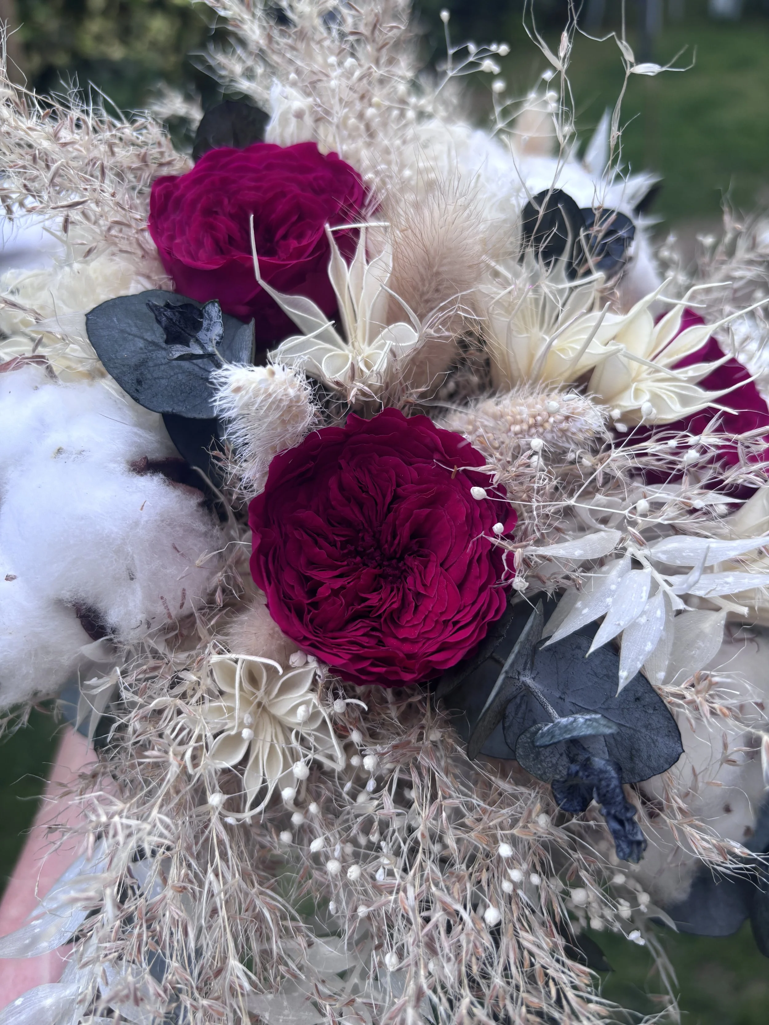 Bouquet de mariée bohème aux nuances bordeaux profond, ivoire et beige naturel, réalisé à partir de fleurs séchées et stabilisées. Cette création florale associe roses stabilisées bordeaux, coton, eucalyptus, graminées aériennes et fleurs texturées p