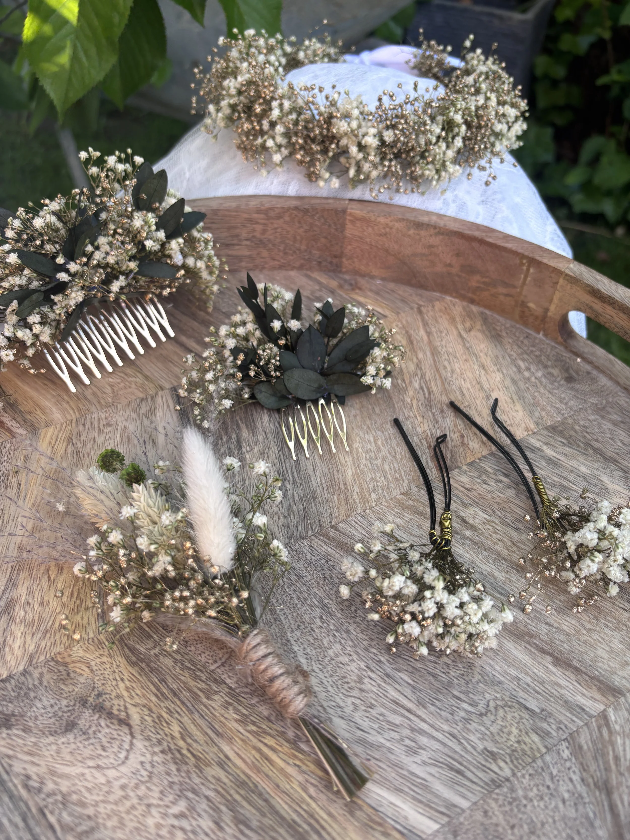 Collection de fleurs de cérémonie avec fleurs blanches, feuillage vert foncé, et accessoires comme des peignes, sur une surface en bois.