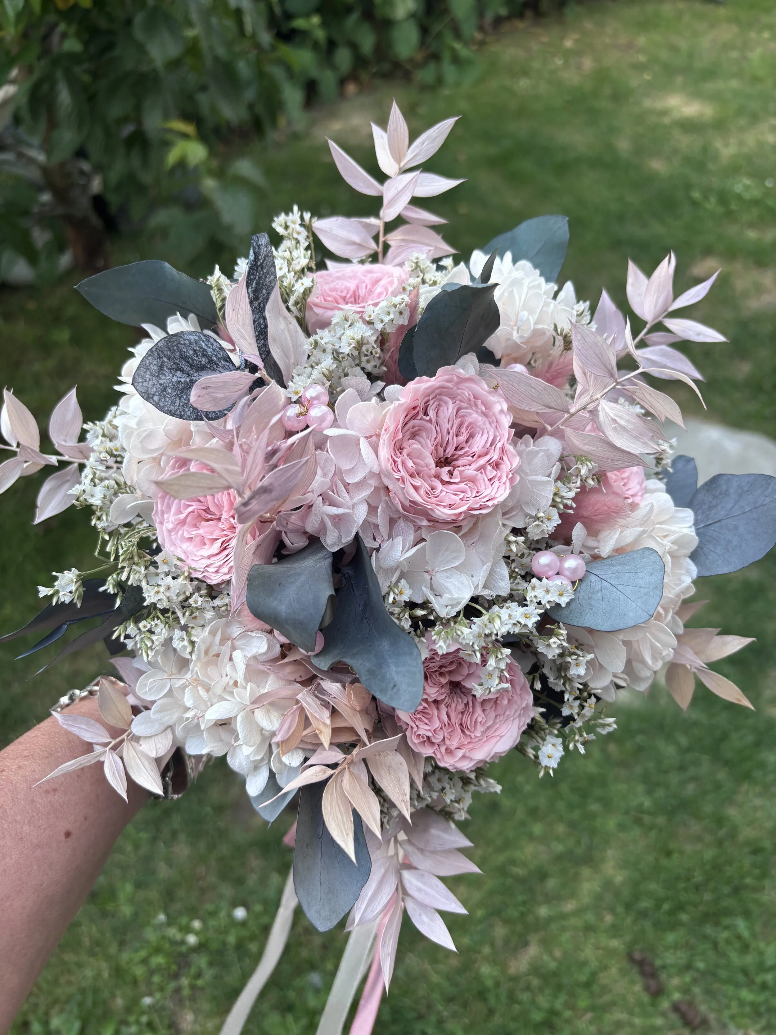 Bouquet de mariée bohème réalisé à la main en fleurs séchées et stabilisées dans des tons rose poudré, blanc et gris bleuté. Cette création florale associe de délicates roses anglaises stabilisées, des hortensias préservés, de l’eucalyptus et des feu