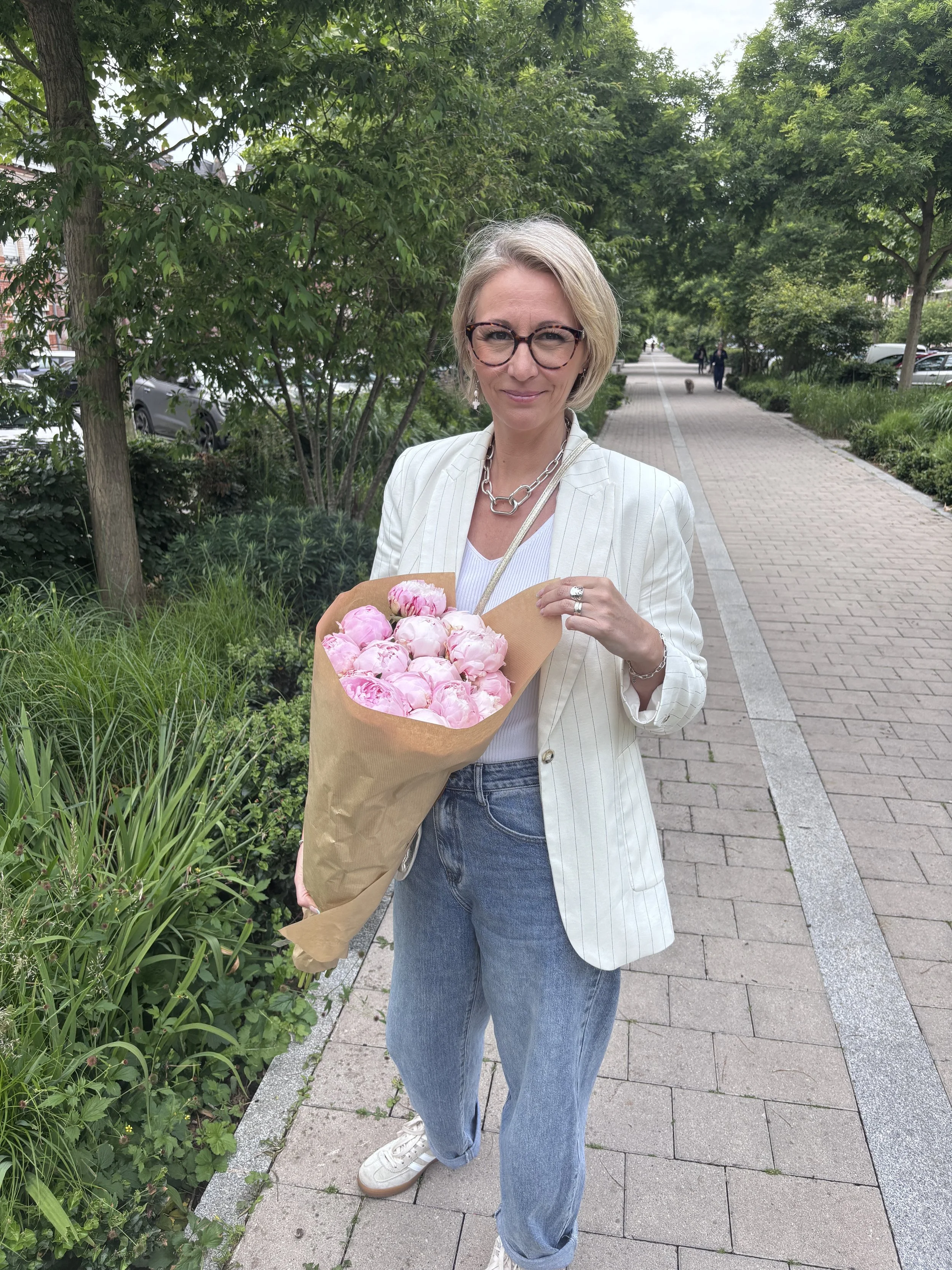 Une femme blonde portant des lunettes, une veste blanche, un t-shirt blanc, et une chaîne en métal, tient un bouquet de roses roses dans un environnement urbain avec des arbres et une passerelle piétonne.