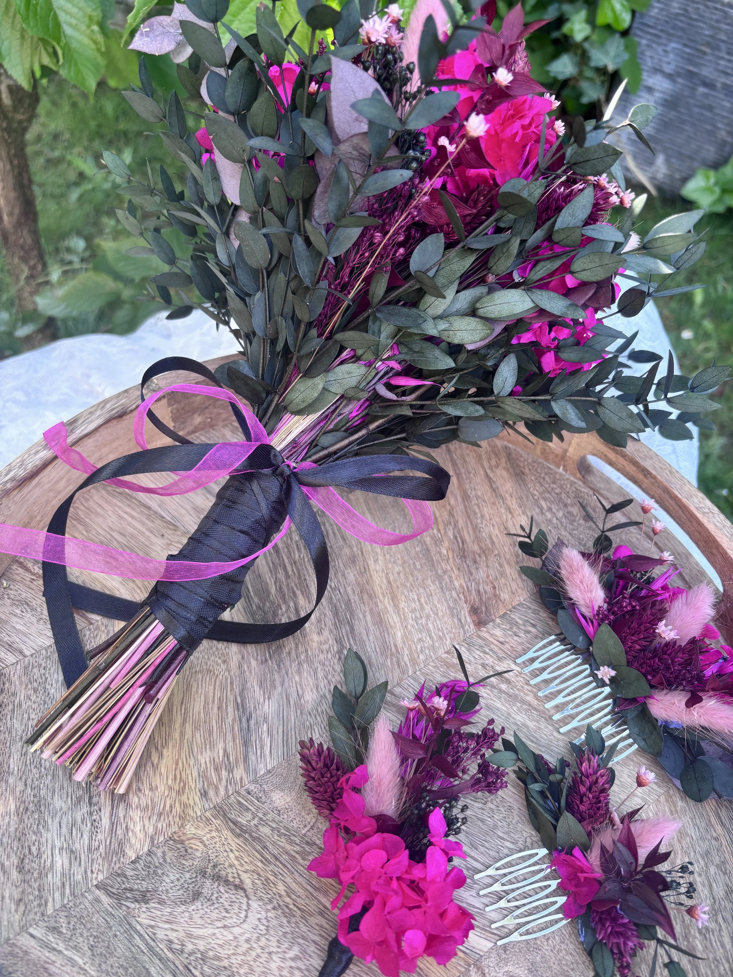 Un bouquet de fleurs roses et violettes avec des feuilles vertes, attaché avec des rubans noirs et roses, posé sur une table en bois.