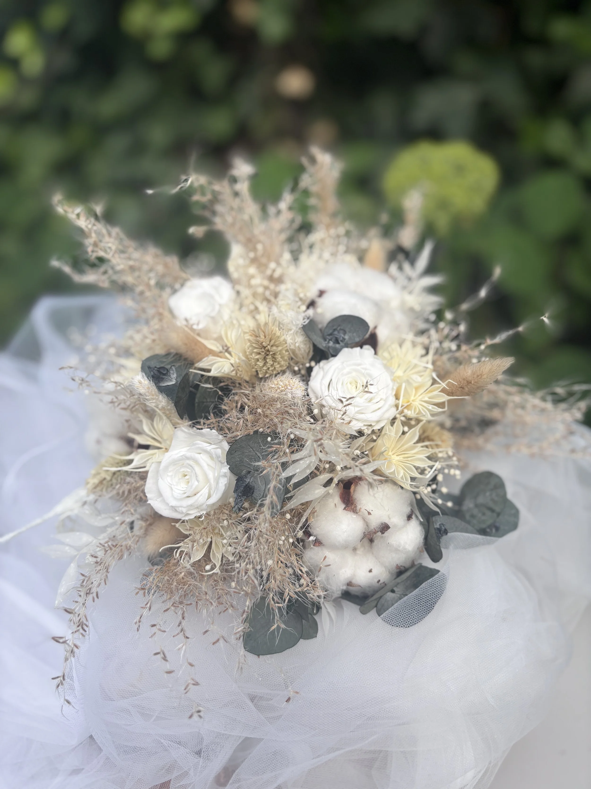 Bouquet de mariée bohème chic réalisé en fleurs séchées et stabilisées dans des nuances de blanc, ivoire, beige naturel et vert eucalyptus. Cette création artisanale associe roses stabilisées, coton, graminées aériennes, eucalyptus et fleurs texturée