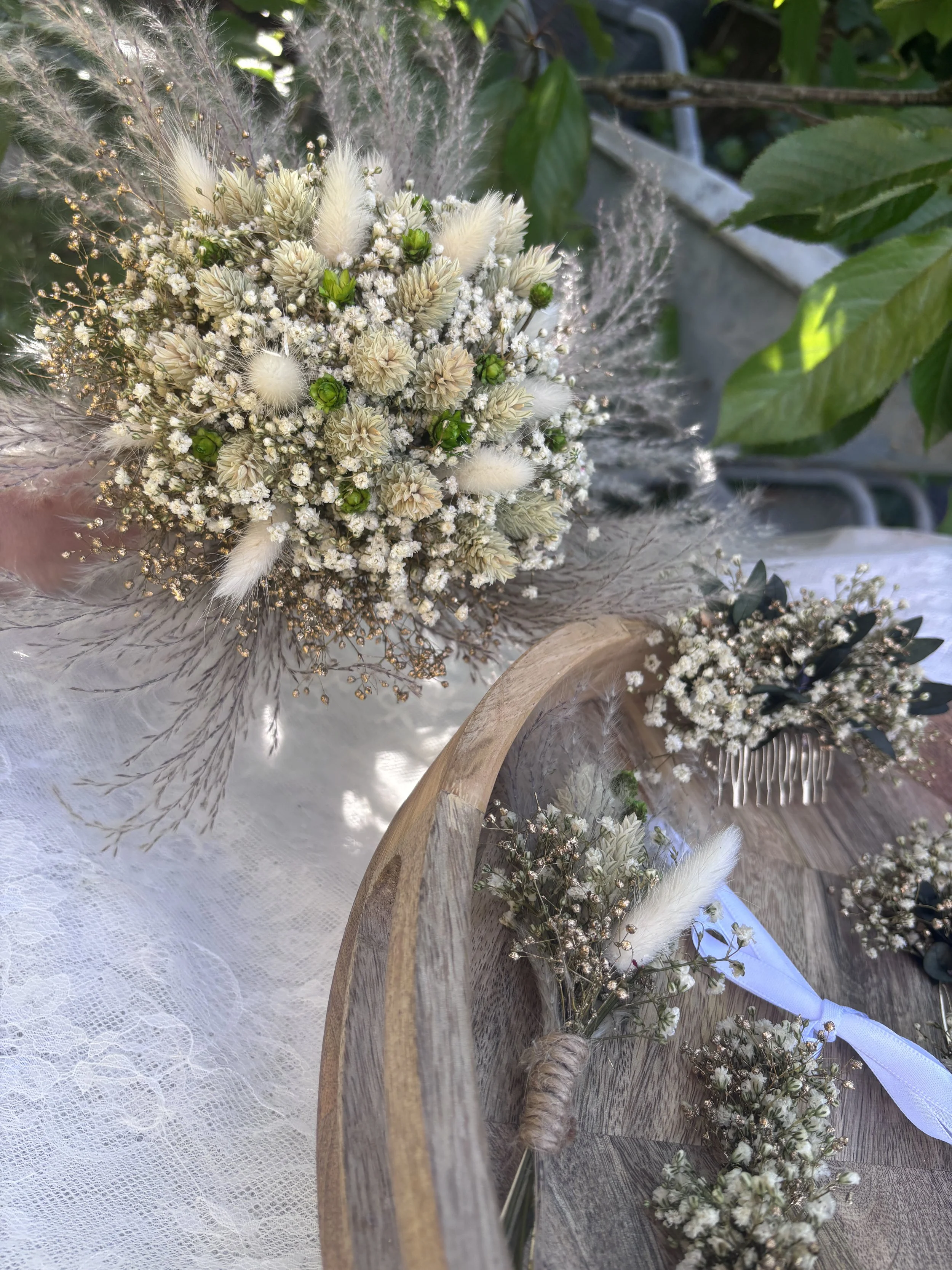 Bouquet de fleurs blanches et vertes avec des plumes et de petits détails dorés, placé sur une table en bois avec une nappe en dentelle blanche.