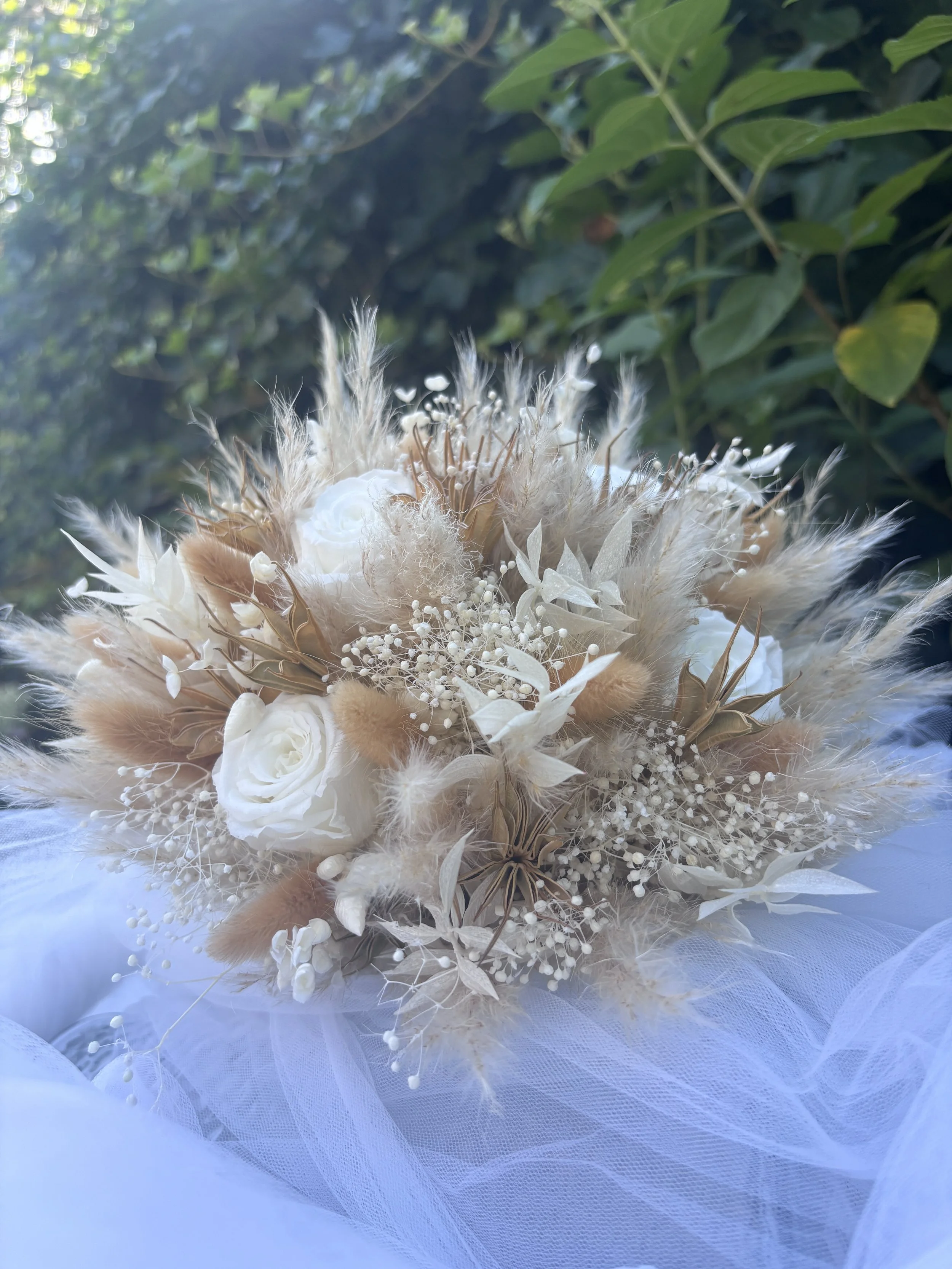 Bouquet de mariée réalisé artisanalement en fleurs séchées et stabilisées. Une création romantique et aérienne mêlant roses blanches, gypsophile, pampas et feuillages naturels dans des nuances ivoire, nude et rose poudré. Création sur mesure par L’At