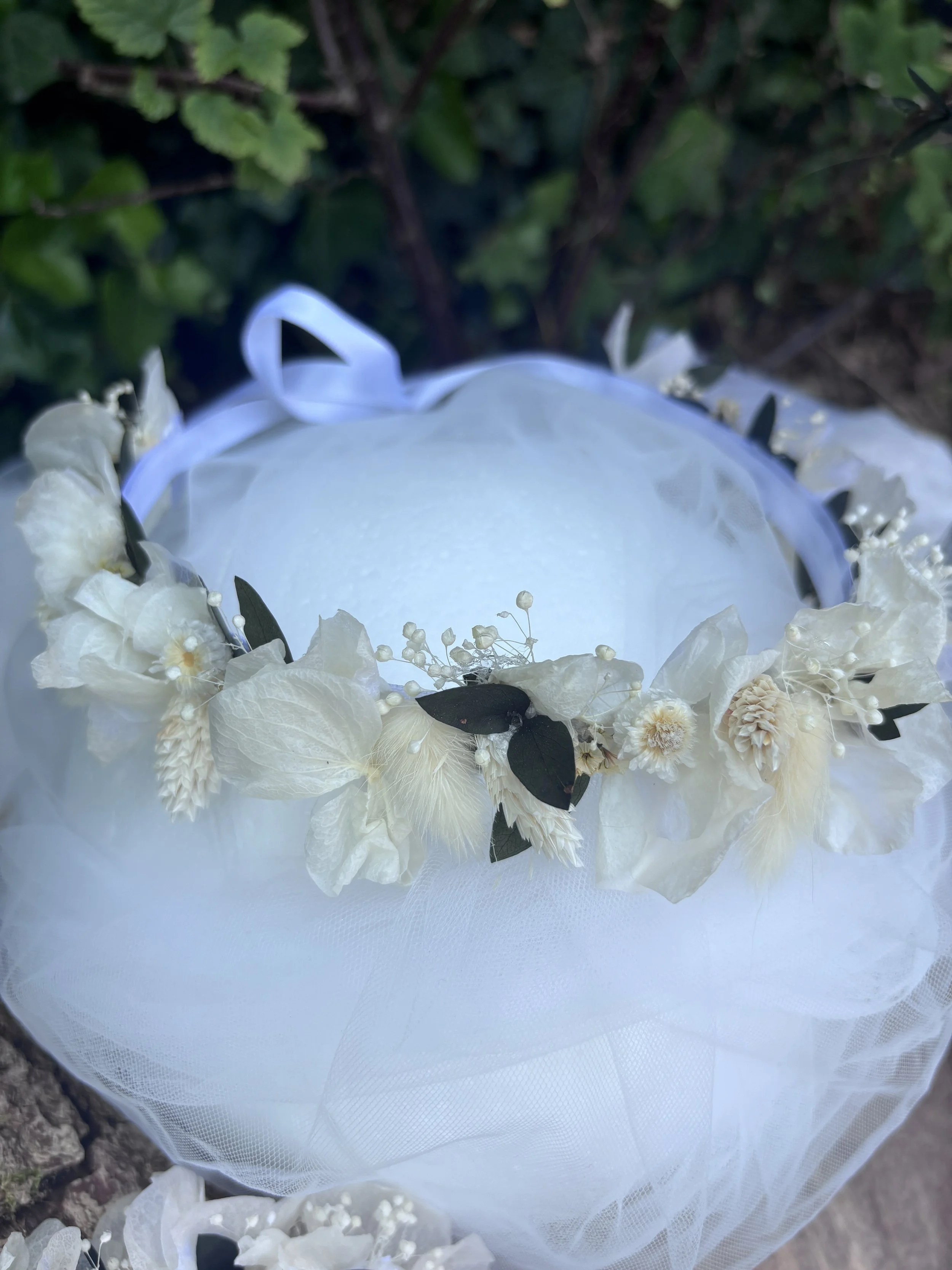 Couronne de fleurs blanches avec des feuilles vertes sur un fond blanc et entourée de tissu en tulle totalement blanc.