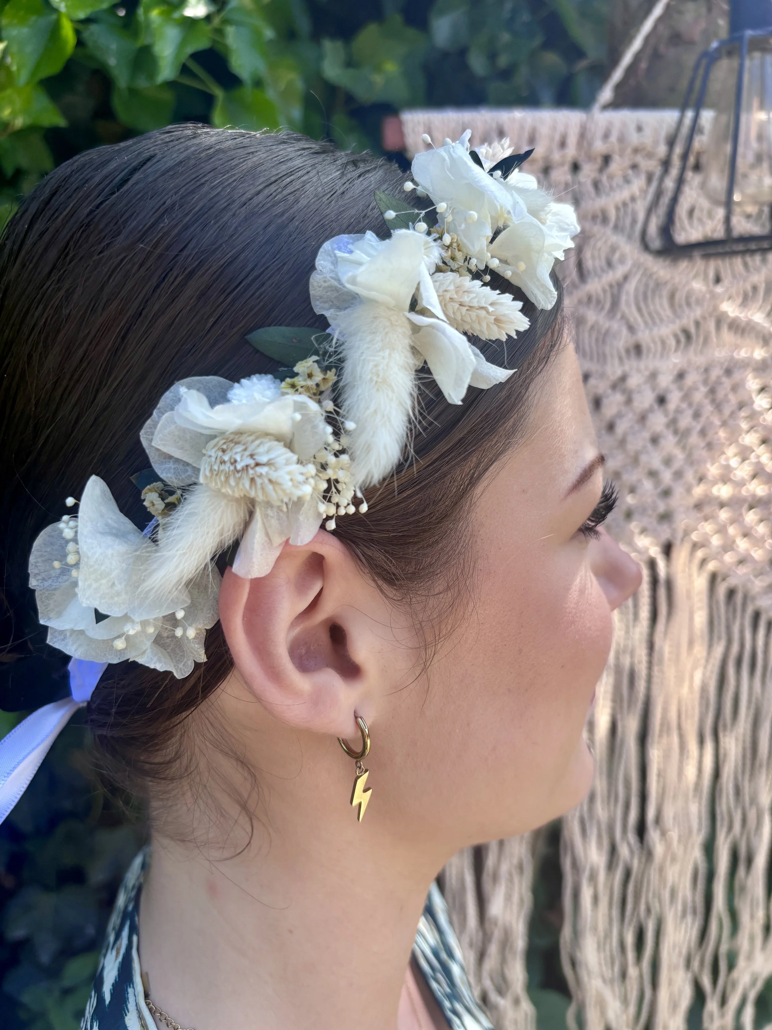 Une jeune femme porte un diadème floral blanc avec des fleurs artificielles et des feuilles, et porte une boucle d'oreille en forme d'éclair en or. La photo est prise du côté droit, avec un fond naturel de végétation et une décoration en corde tressé