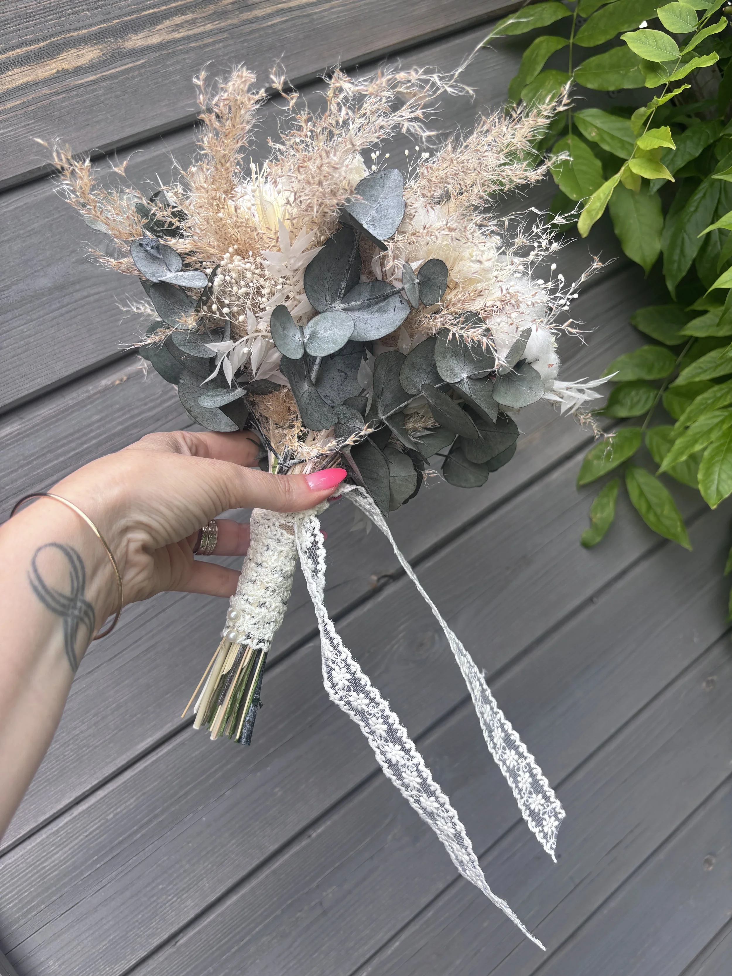 Bouquet de mariée bohème chic réalisé en fleurs séchées et stabilisées dans des nuances de blanc, ivoire, beige naturel et vert eucalyptus. Cette création artisanale associe roses stabilisées, coton, graminées aériennes, eucalyptus et fleurs texturée
