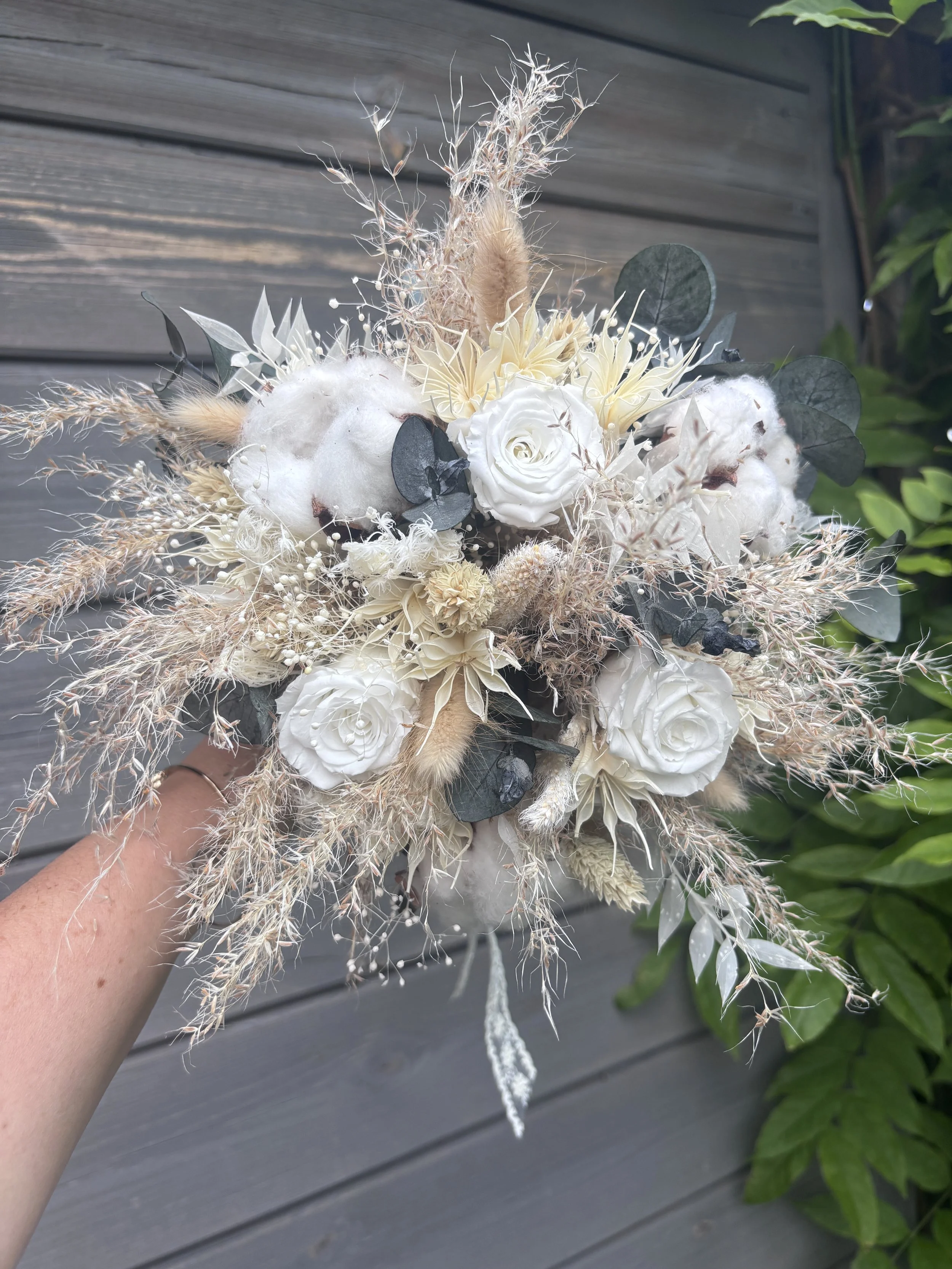 Bouquet de mariée bohème chic réalisé en fleurs séchées et stabilisées dans des nuances de blanc, ivoire, beige naturel et vert eucalyptus. Cette création artisanale associe roses stabilisées, coton, graminées aériennes, eucalyptus et fleurs texturée