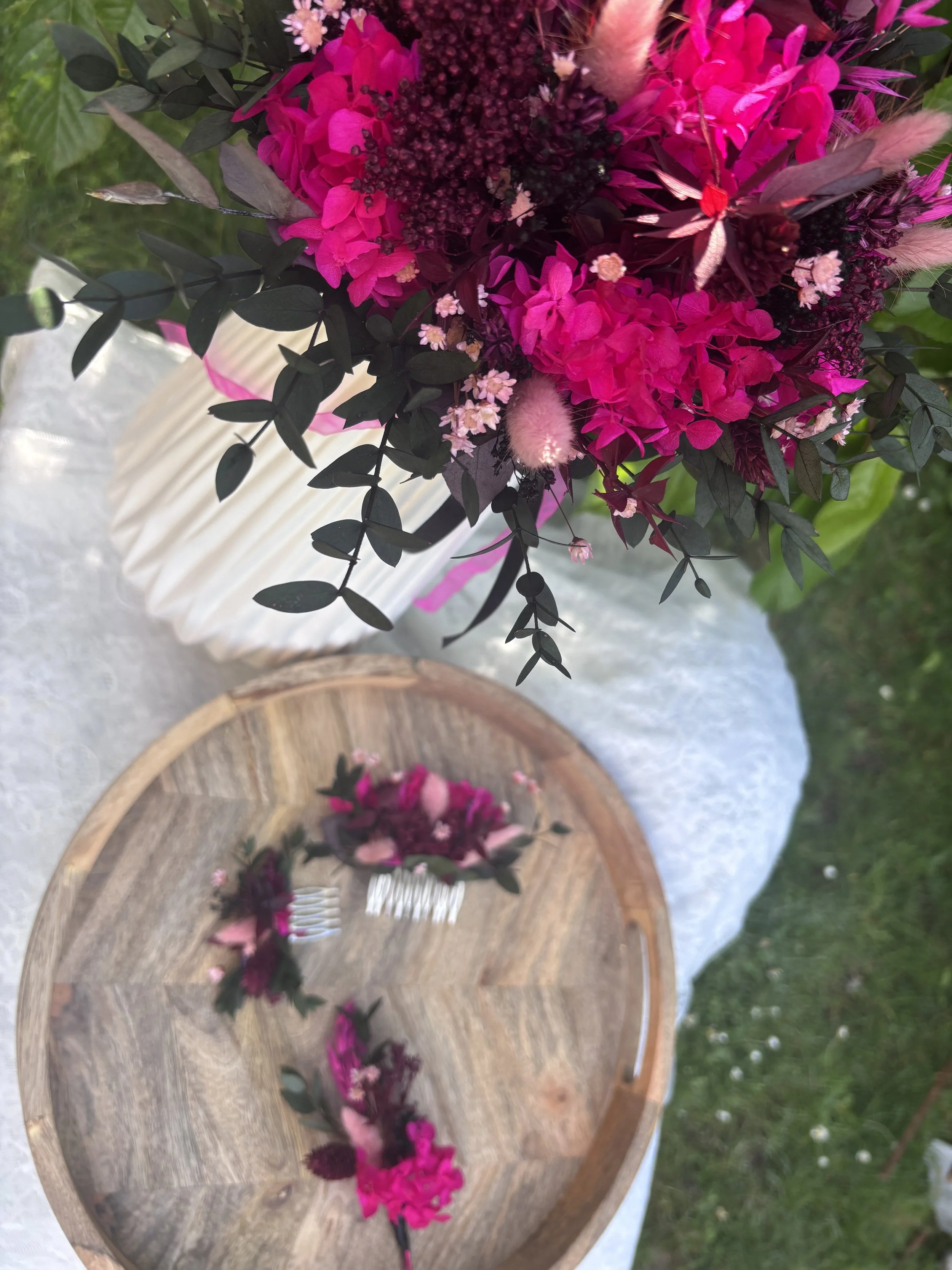 Un bouquet de fleurs violettes, roses et blanches dans un vase blanc. Sur une table en bois, il y a aussi des vases avec des fleurs, et une fourchette avec des fleurs roses et noires, sur un fond vert.