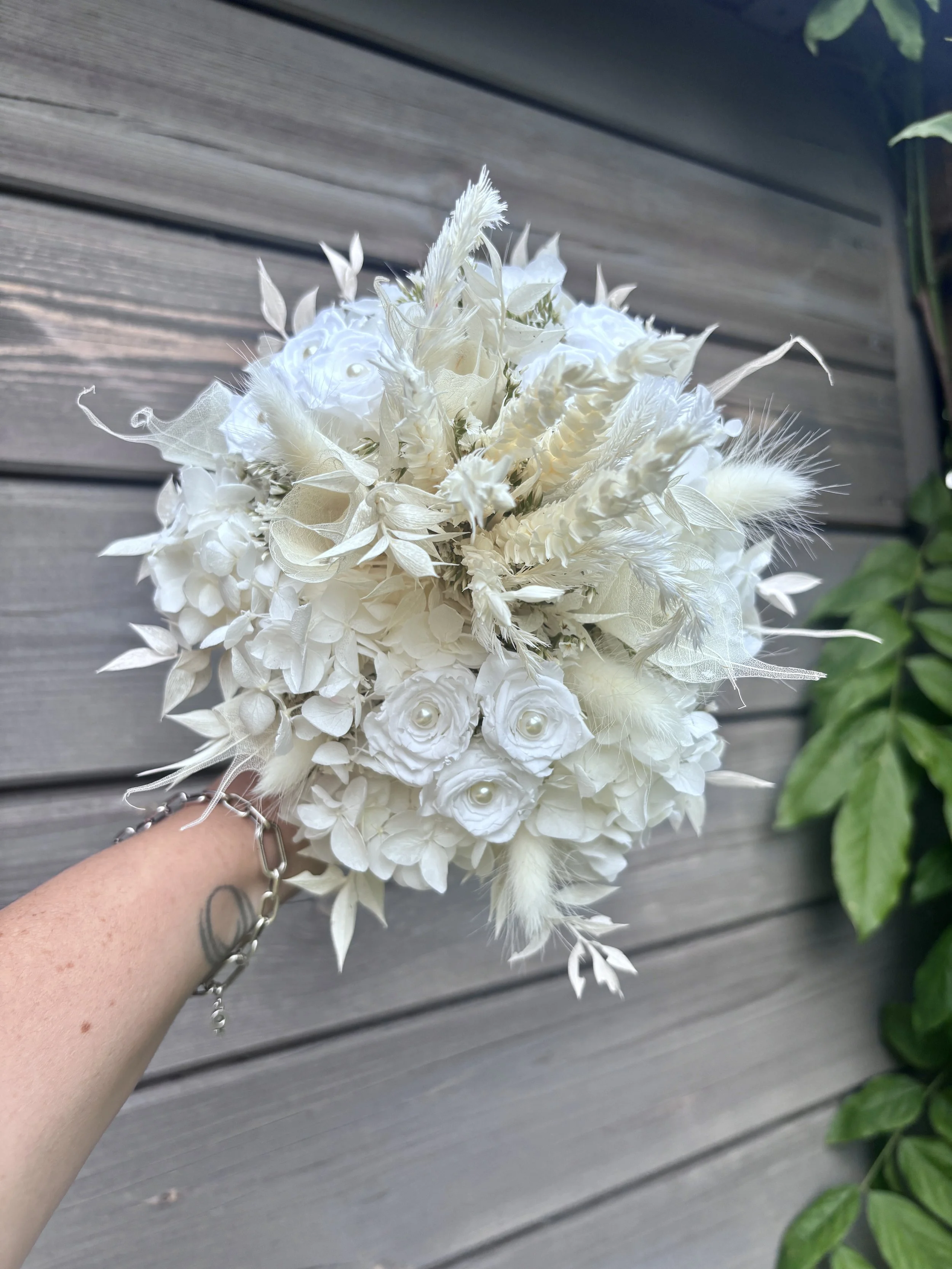 Bouquet de mariée en fleurs séchées et stabilisées blanches, réalisé artisanalement dans un esprit bohème chic et romantique. Cette création florale associe hortensias stabilisés, roses éternelles ornées de perles, arums délicats, feuillages blanchis