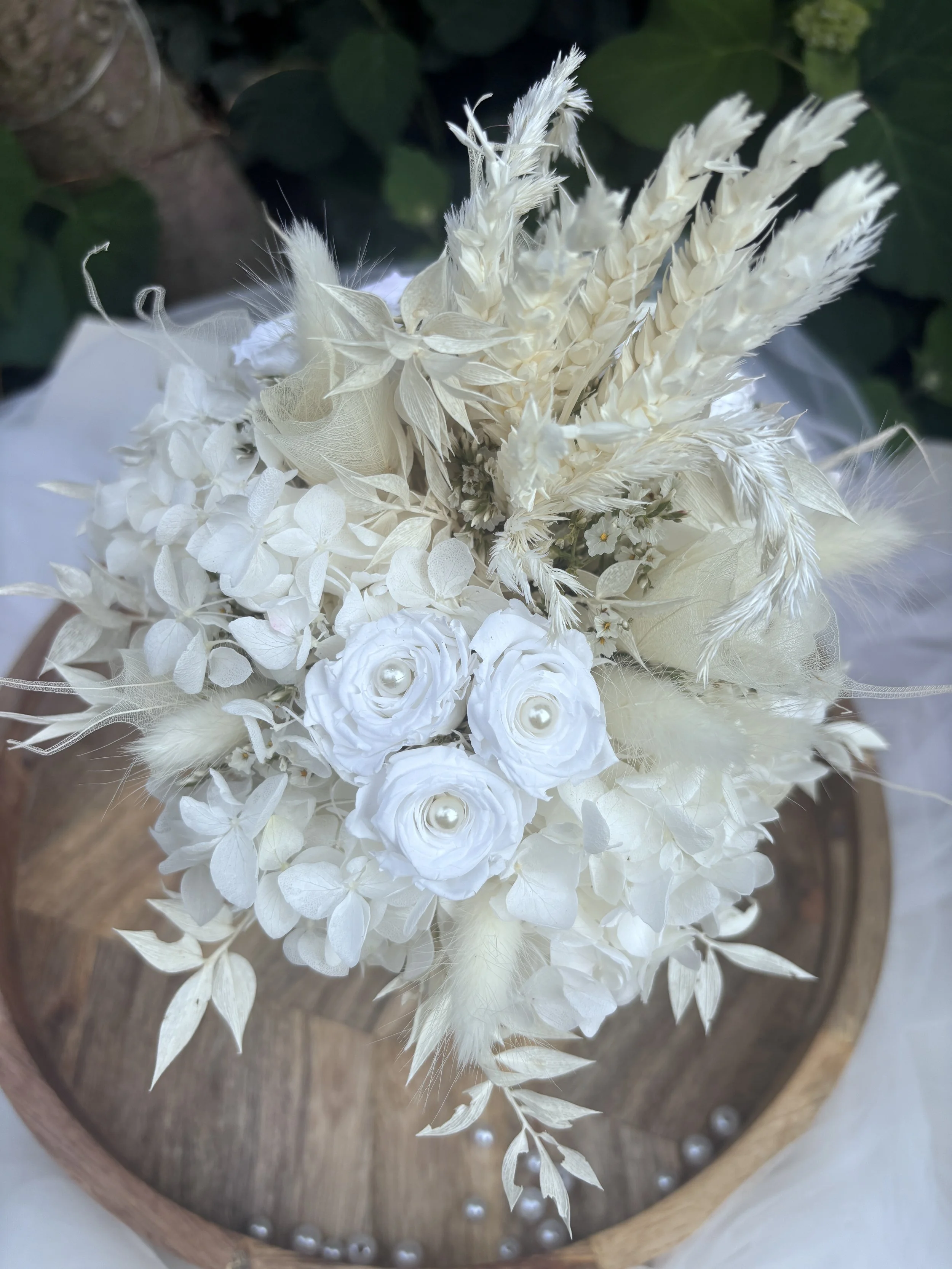 Bouquet de mariée en fleurs séchées et stabilisées blanches, réalisé artisanalement dans un esprit bohème chic et romantique. Cette création florale associe hortensias stabilisés, roses éternelles ornées de perles, arums délicats, feuillages blanchis