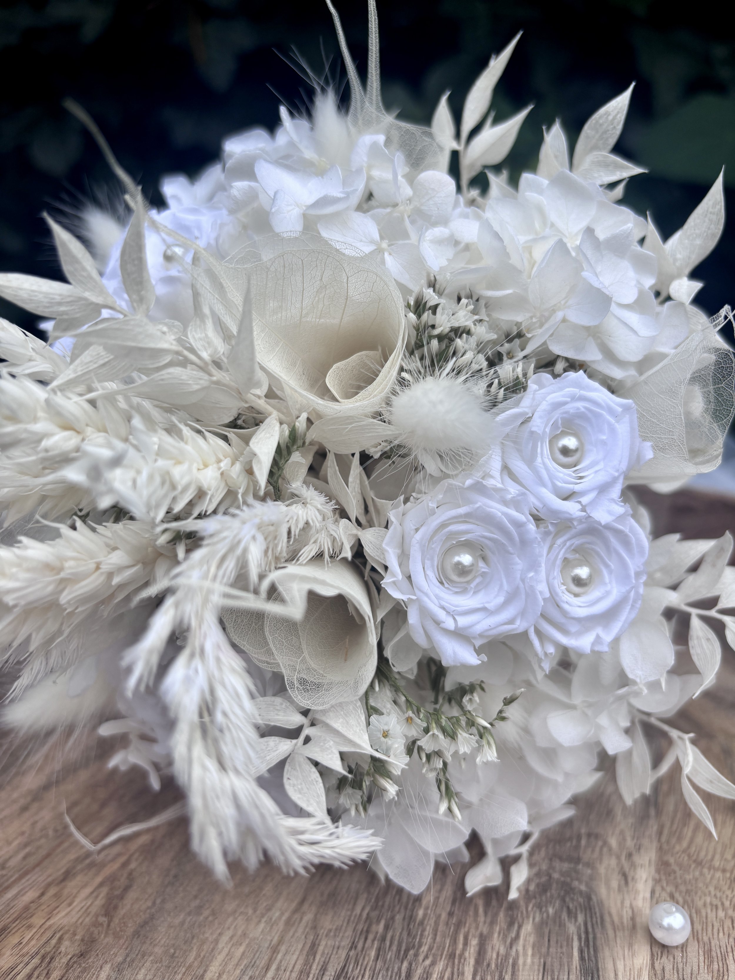 Bouquet de mariée en fleurs séchées et stabilisées blanches, réalisé artisanalement dans un esprit bohème chic et romantique. Cette création florale associe hortensias stabilisés, roses éternelles ornées de perles, arums délicats, feuillages blanchis