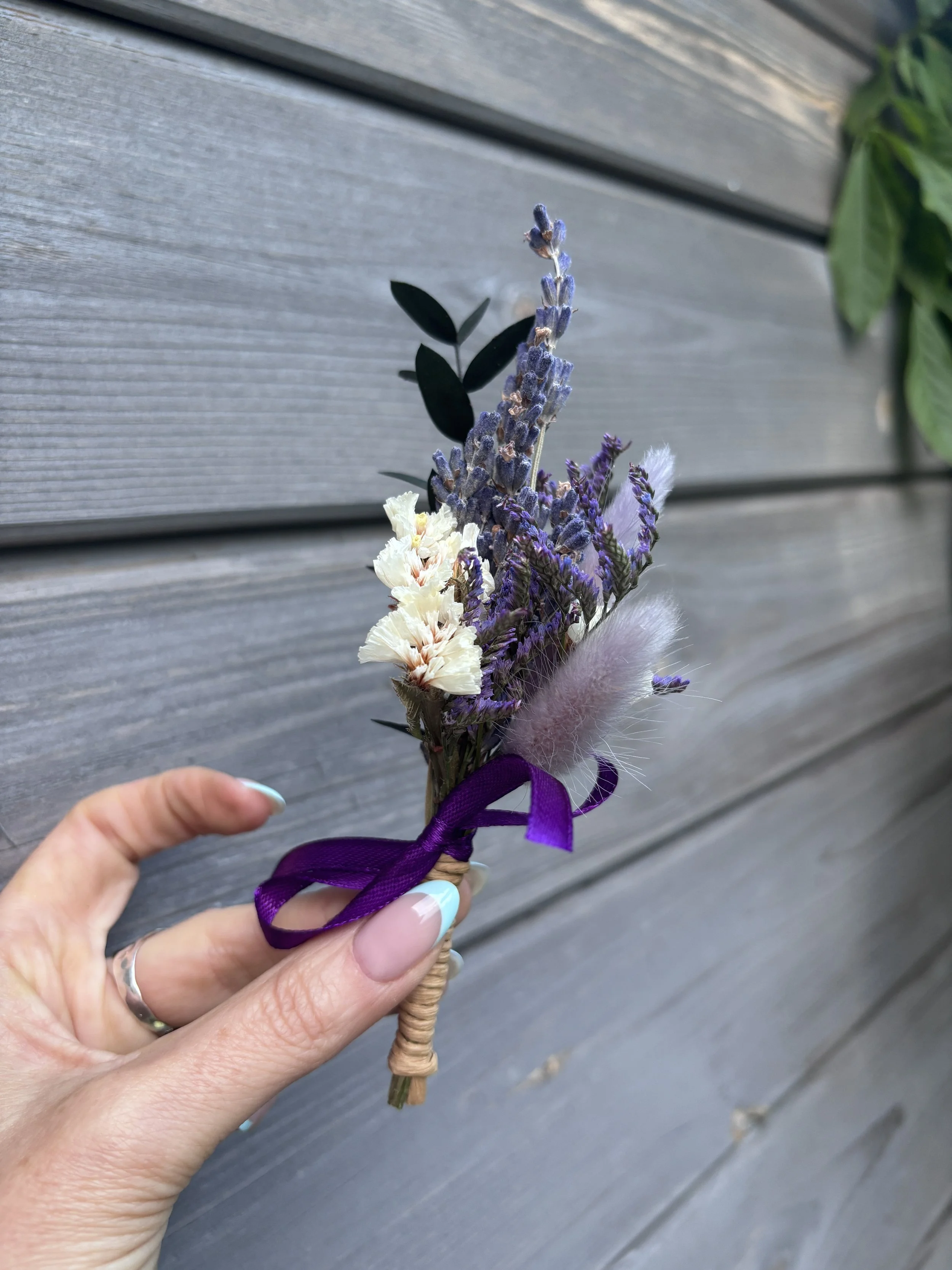 Mains tenant un petit bouquet de fleurs séchées avec une lavande violette, des fleurs blanches, et une touche de rose, attaché avec un ruban violet, devant un fond de planche en bois grise.
