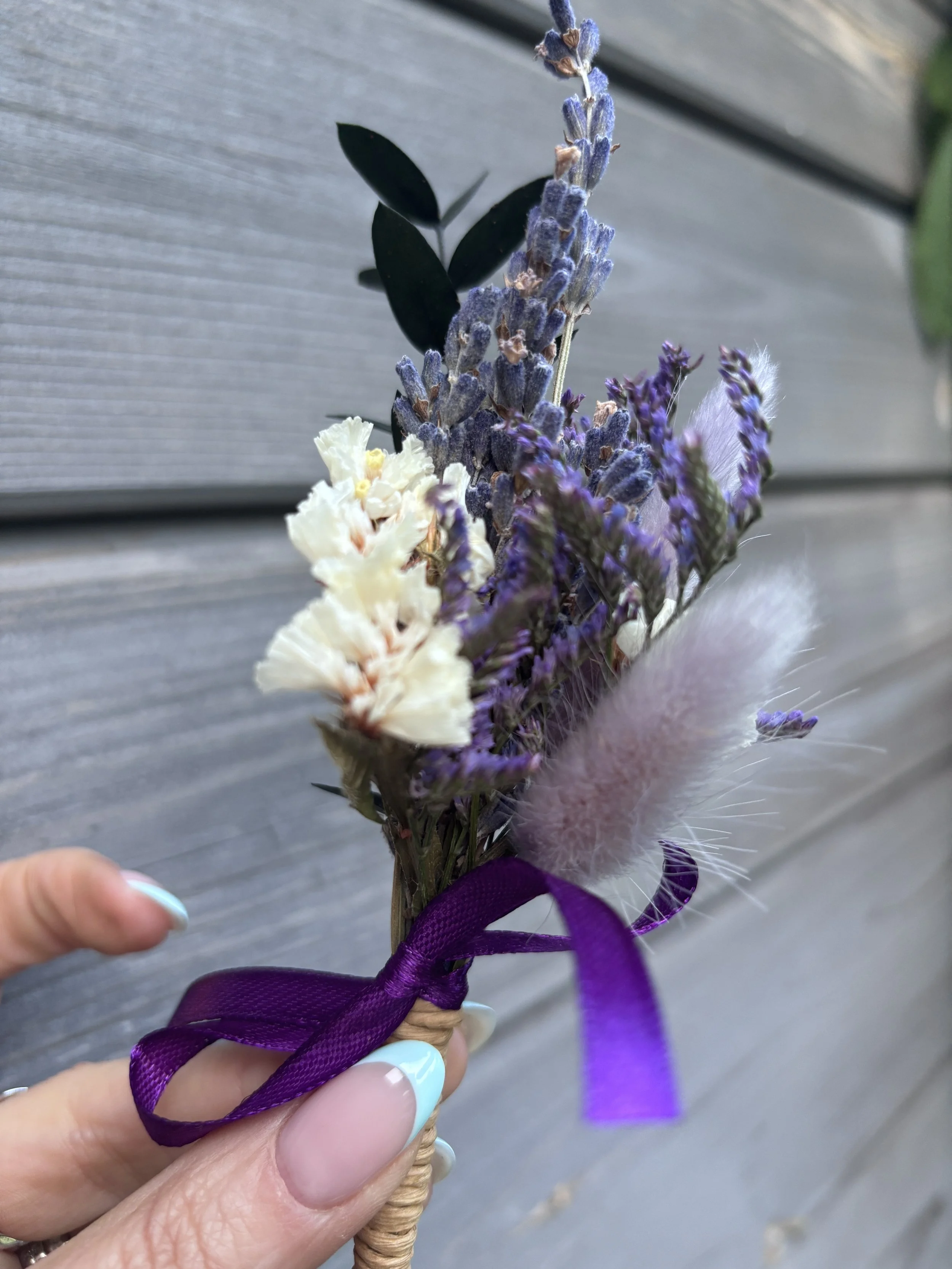 Main tenant une petite décoration florale composée de lavande violette et de fleurs blanches, attachée avec un ruban violet, proche d'une main avec des ongles peints en blanc et bleu pastel, contre un fond en bois.