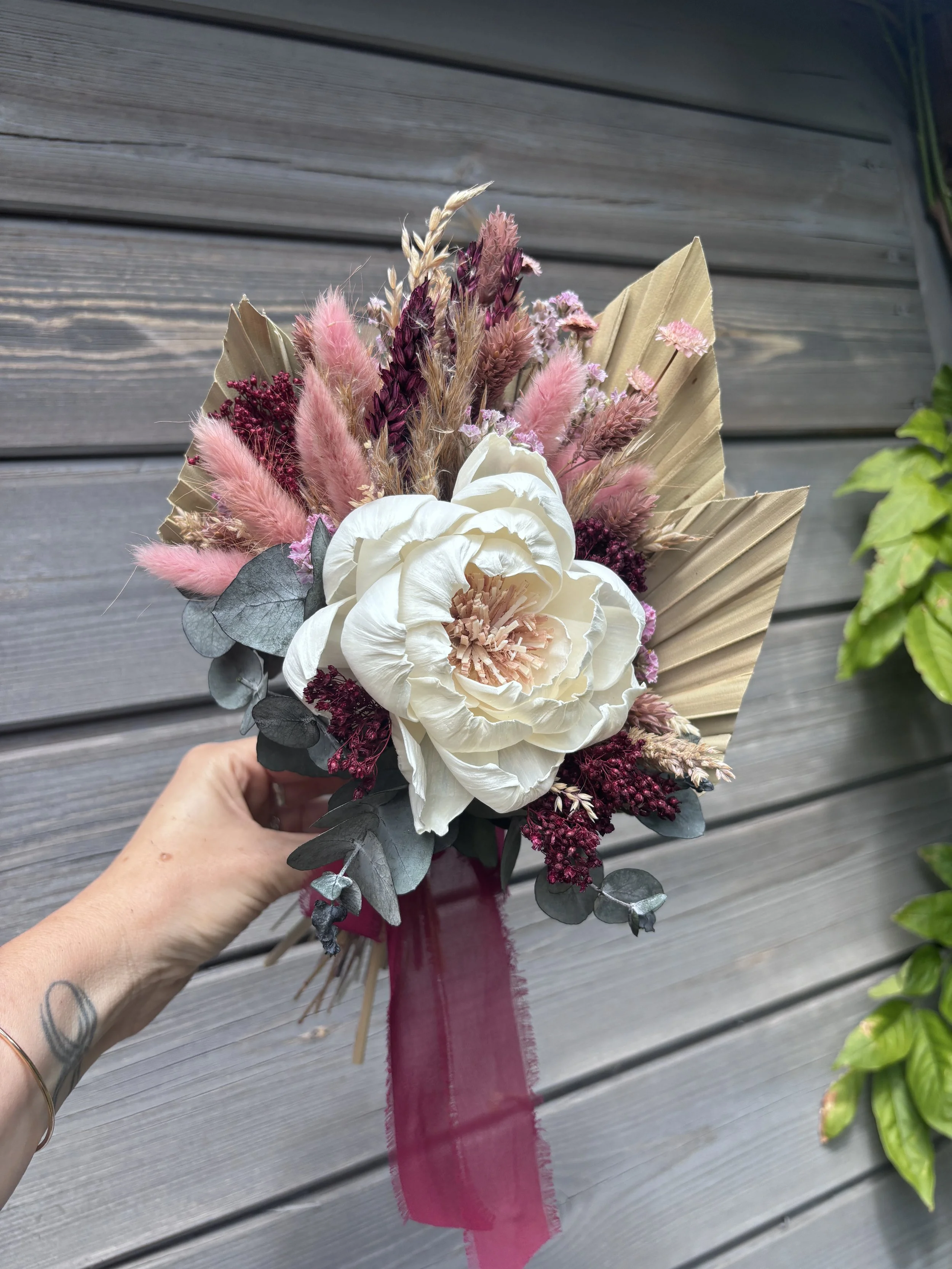 Bouquet de mariée en fleurs séchées et stabilisées, composé d’une grande pivoine ivoire en bois de Sola, d’eucalyptus, de lagurus rose poudré, de fleurs bordeaux et de palmes naturelles. Un bouquet romantique et durable, idéal pour un mariage bohème 